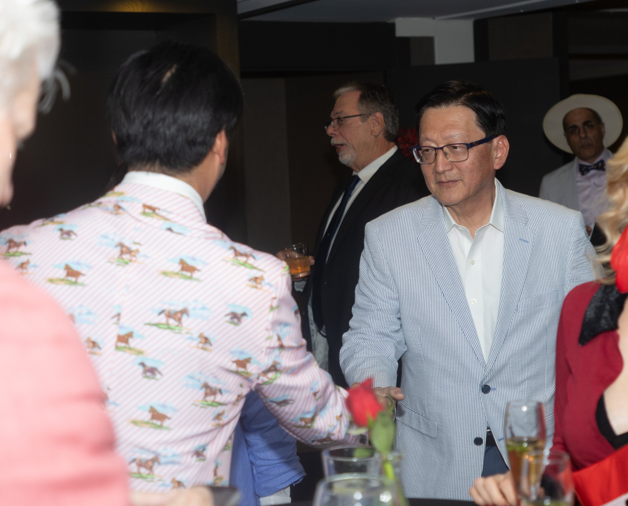 A group of people at a social gathering or party, engaging in conversation. Some are holding drinks. The focus is on a man with glasses and a light striped blazer.