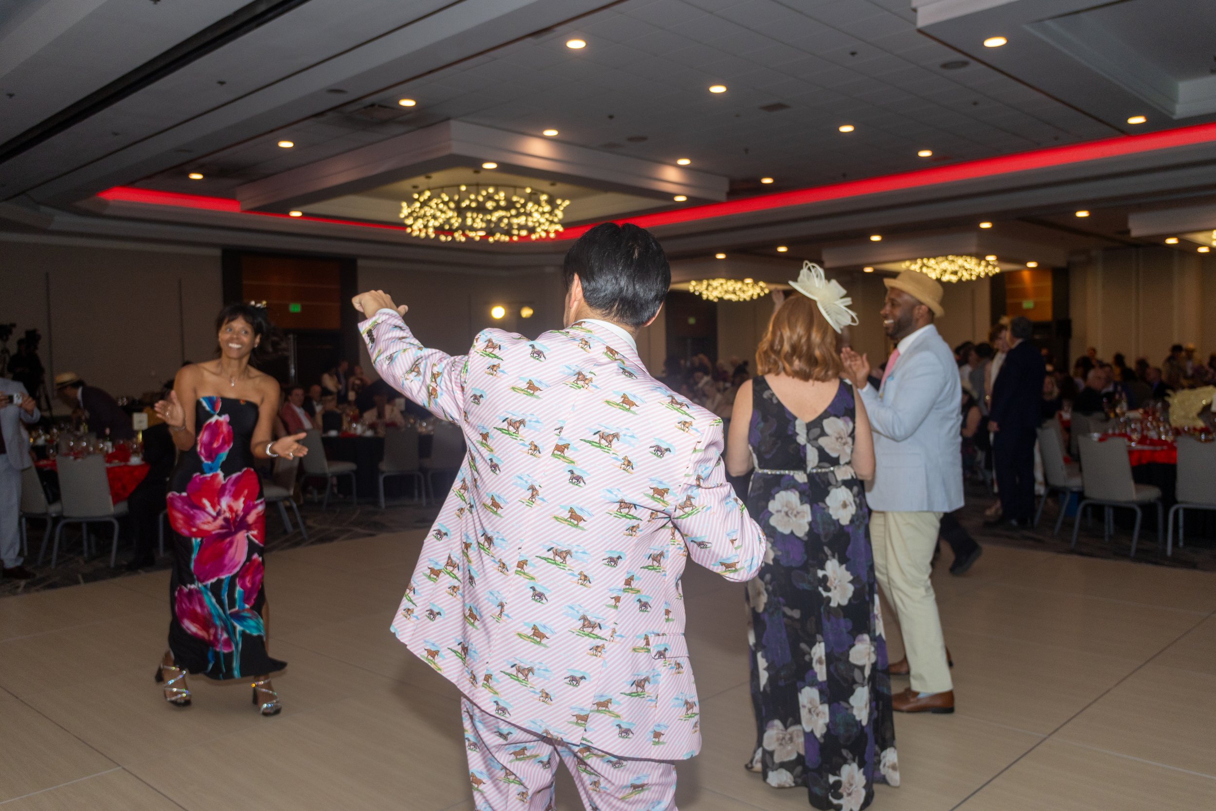 People dancing at a formal indoor event, some in colorful and patterned clothing, with a decorated ceiling and round tables in the background.