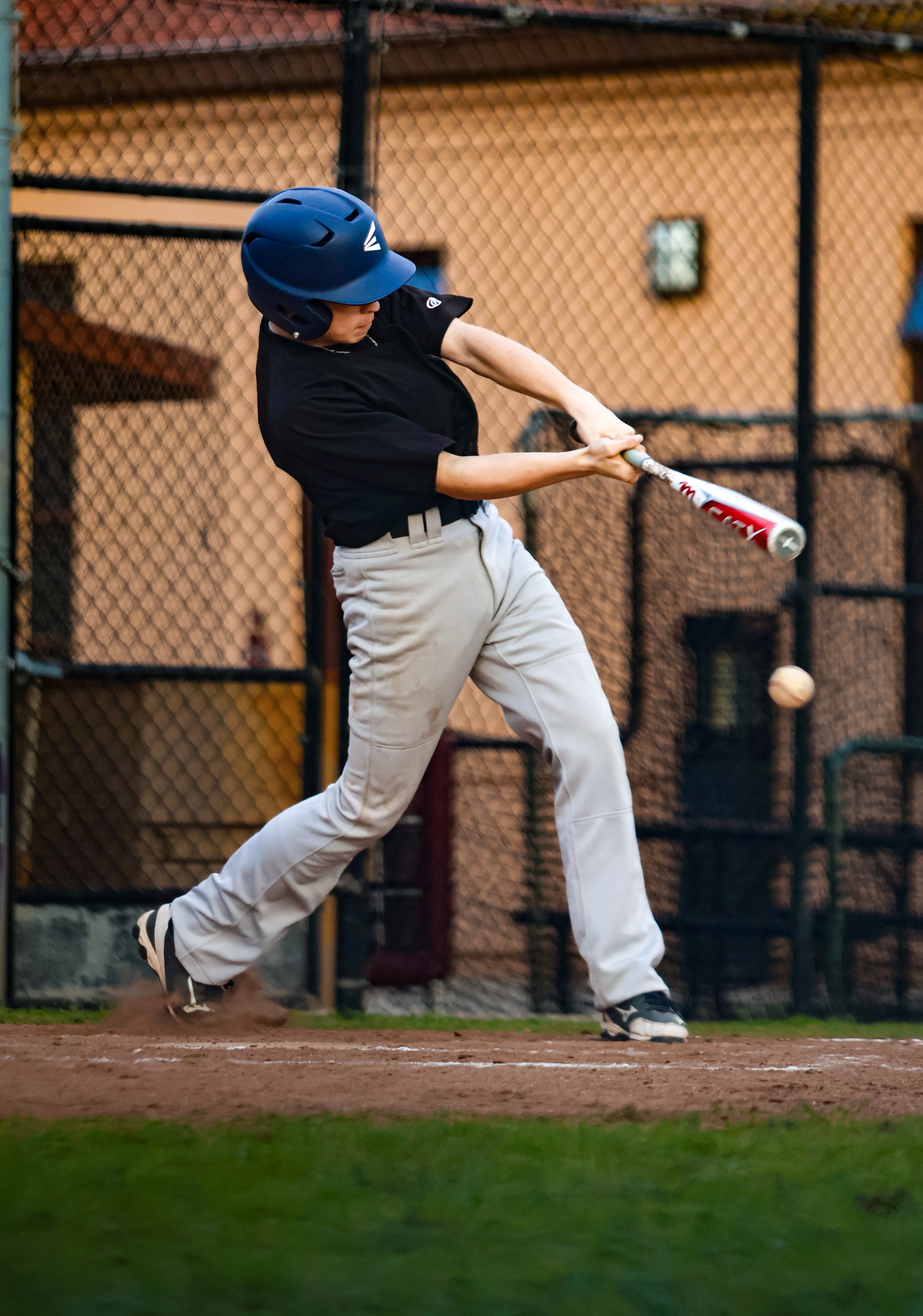 A baseball player wearing a dark helmet, black shirt, and beige pants is swinging a bat during a game on a dirt field with a chain-link fence in the background.
