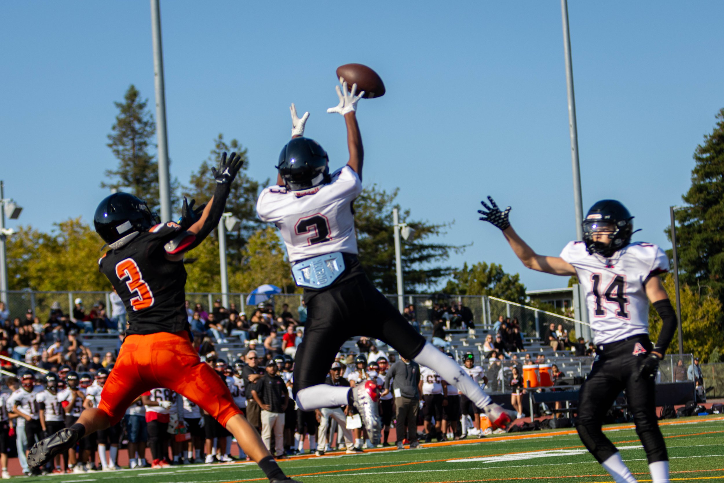 A football player in white uniform jumping to catch a football while two players in black and orange attempt to block him during a game with a crowd in the background.