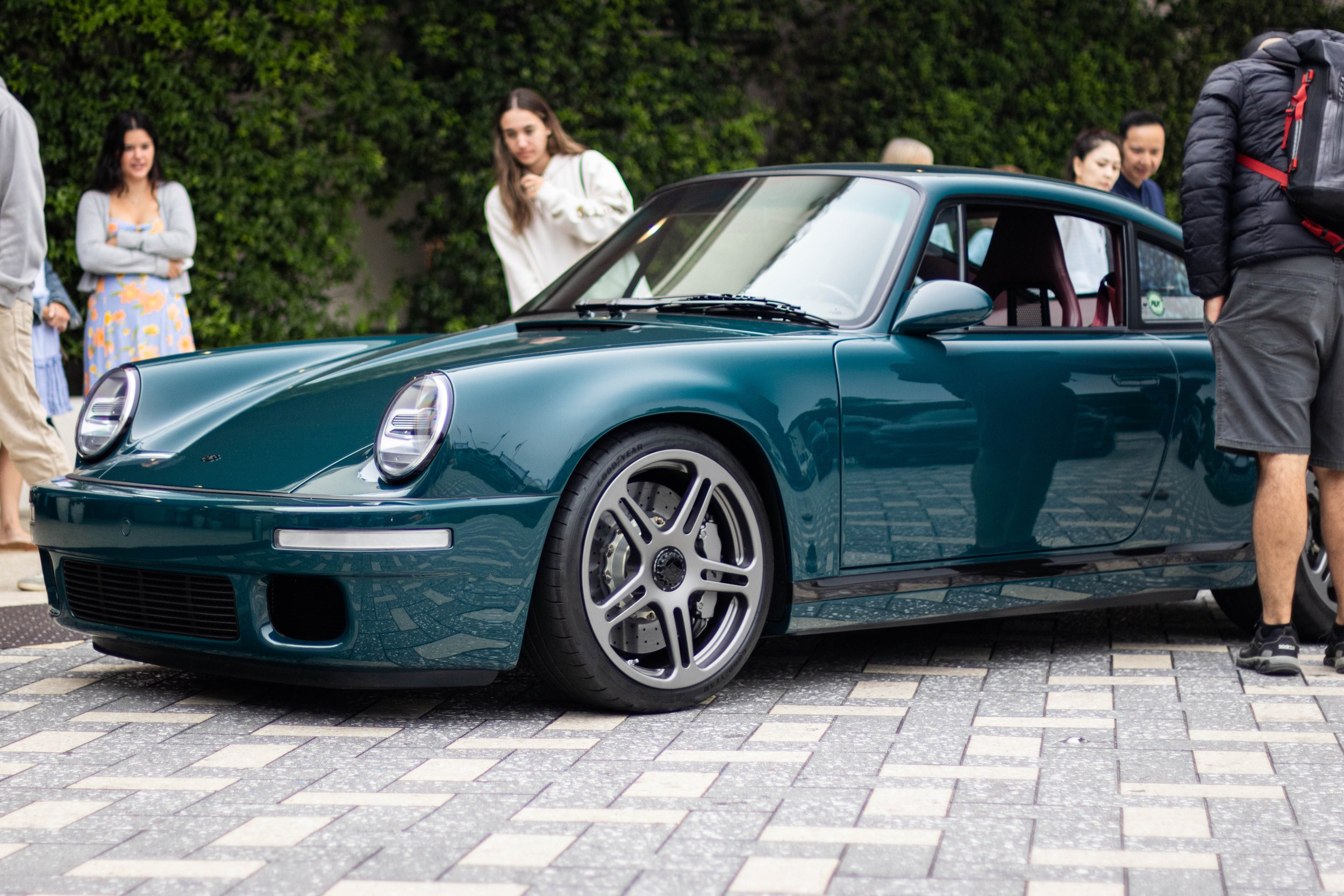 A teal Porsche sports car parked on a cobblestone street with several people around, some looking at the car and others engaged in conversation, with greenery in the background.