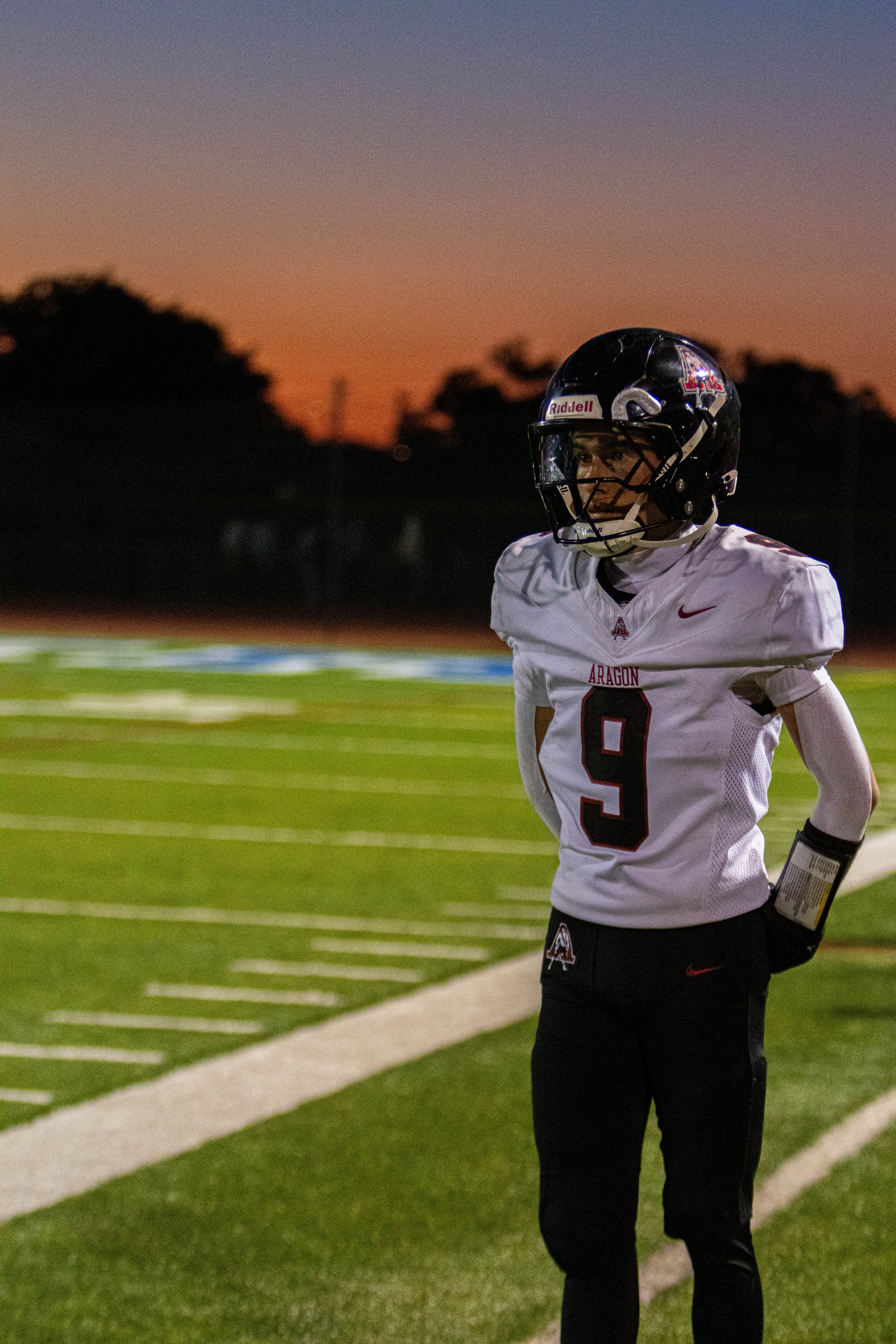 A football player wearing a white jersey with the number 9 and a helmet, standing on the sideline of a football field during sunset.