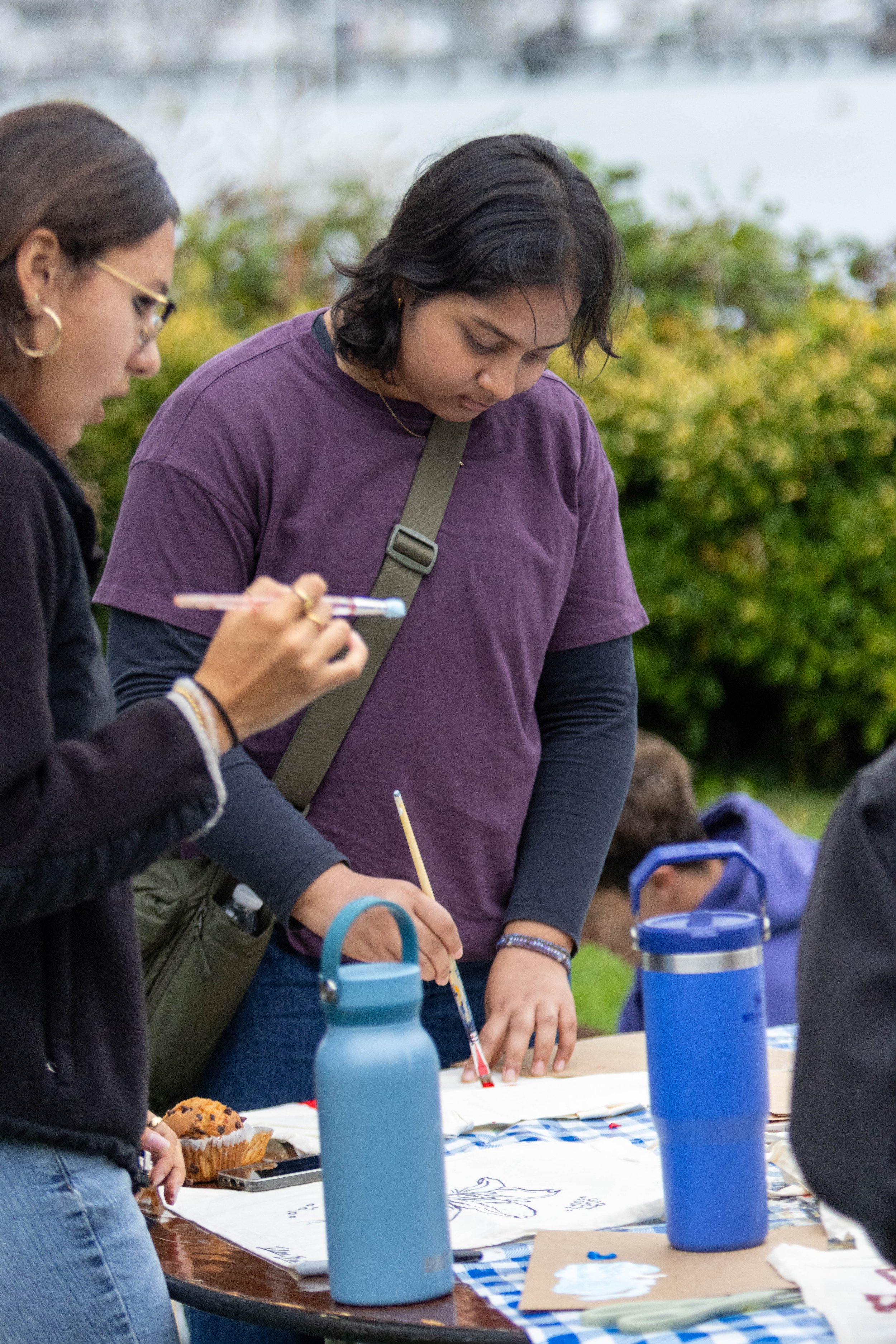 People gathered around a table outdoors, engaging in an art activity with paper, paint, and brushes, with water bottles and snacks on the table, greenery in the background, during daytime.