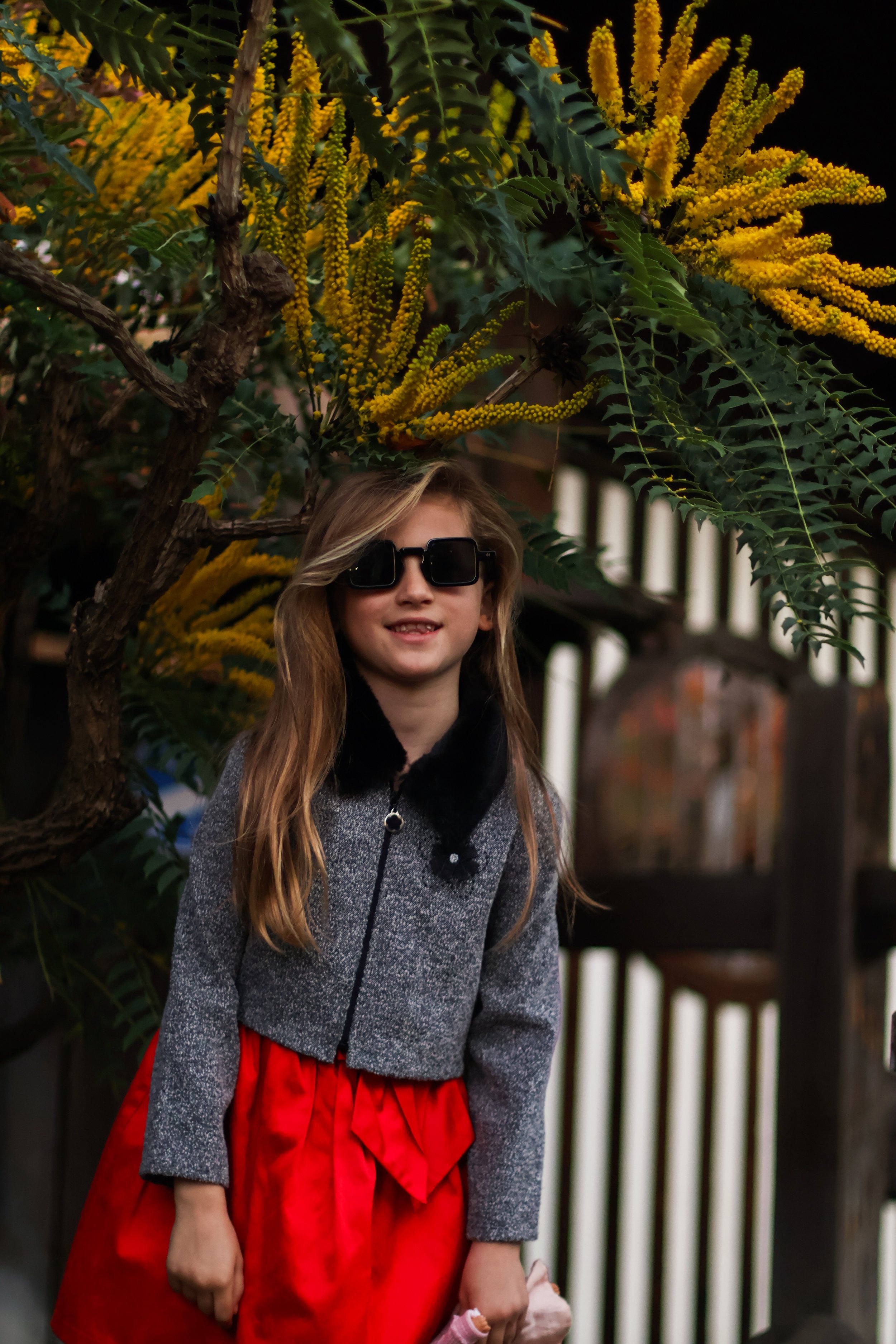 A girl with long blonde hair wearing black sunglasses, a gray coat with black fur collar, and a red skirt, standing outdoors in front of a holly tree with yellow flowers and green leaves at night.