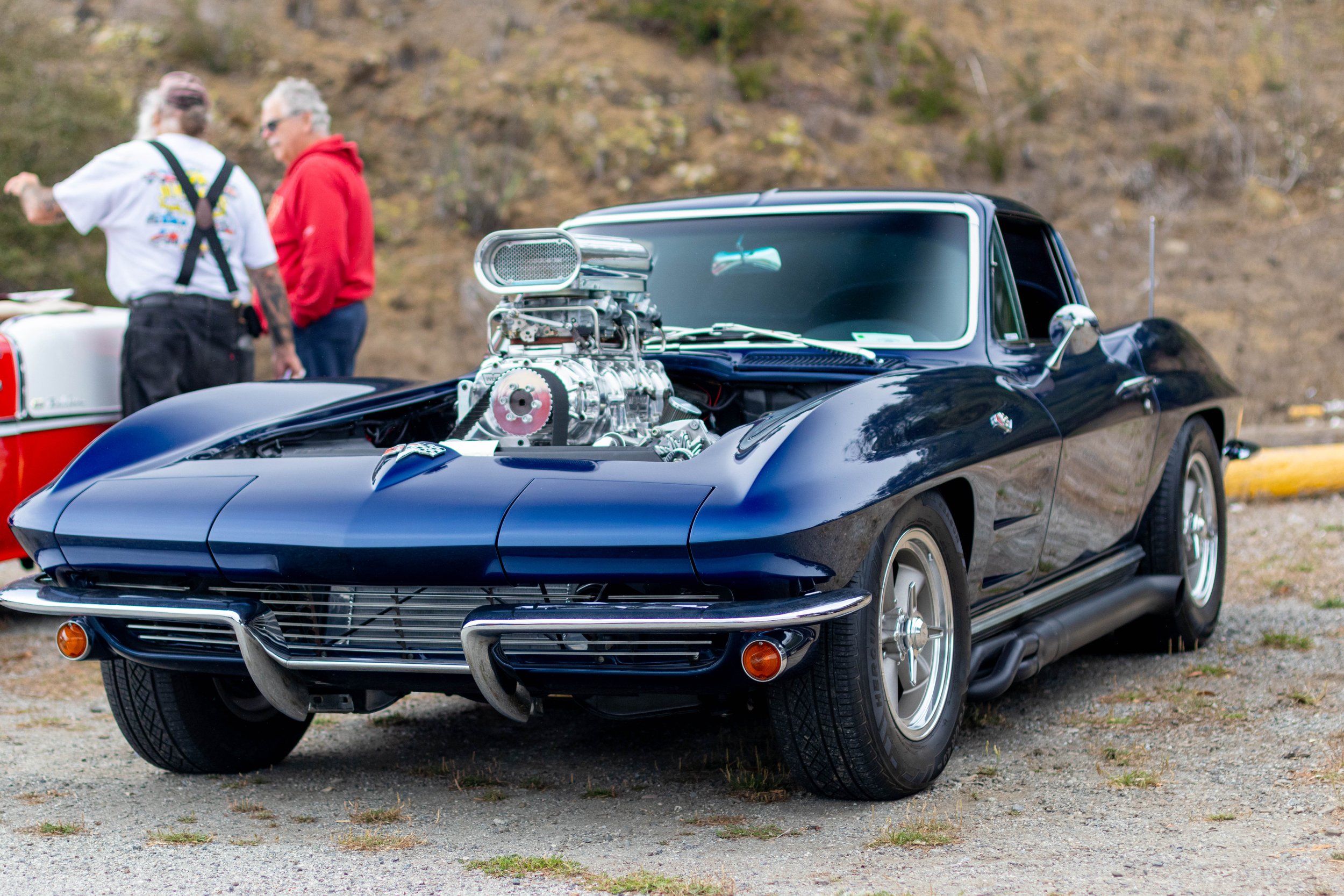 A black classic car with a large chrome engine protruding from the hood, parked outdoors with a rocky hillside in the background. Two men are standing behind the car, one in a white shirt and another in a red hoodie, engaged in conversation.
