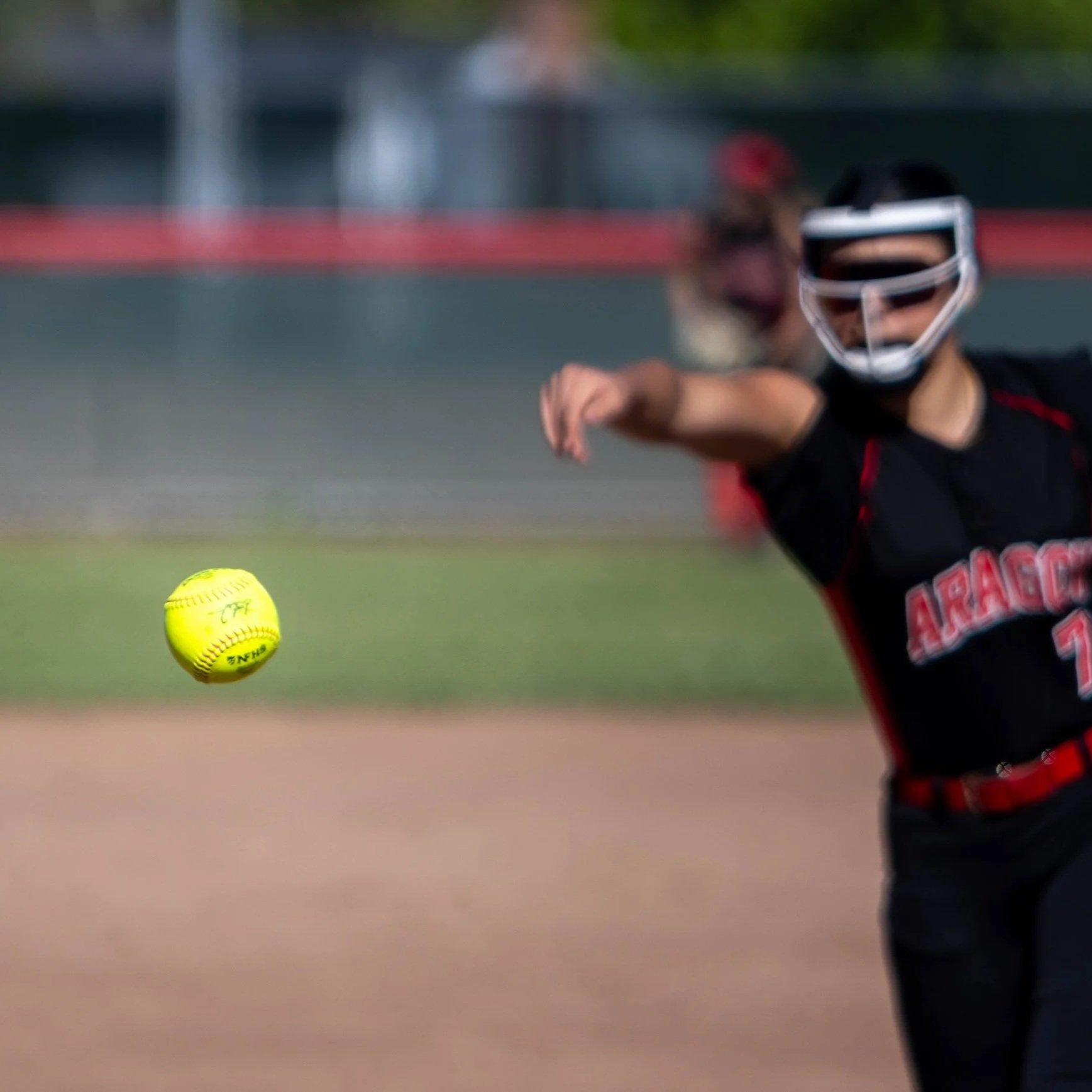 A softball player in black and red uniform preparing to catch a yellow softball, with a blurred background of a baseball field.