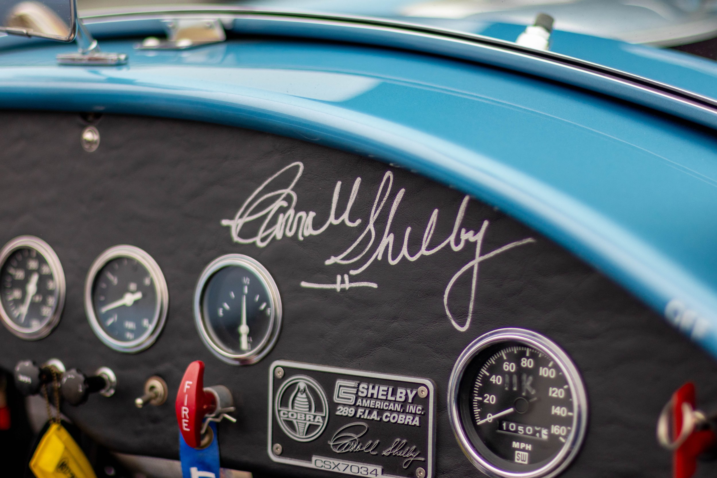 Close-up of a classic car dashboard featuring analog gauges, a signature plate reading 'Russell Shelby,' and various racing stickers and tags.