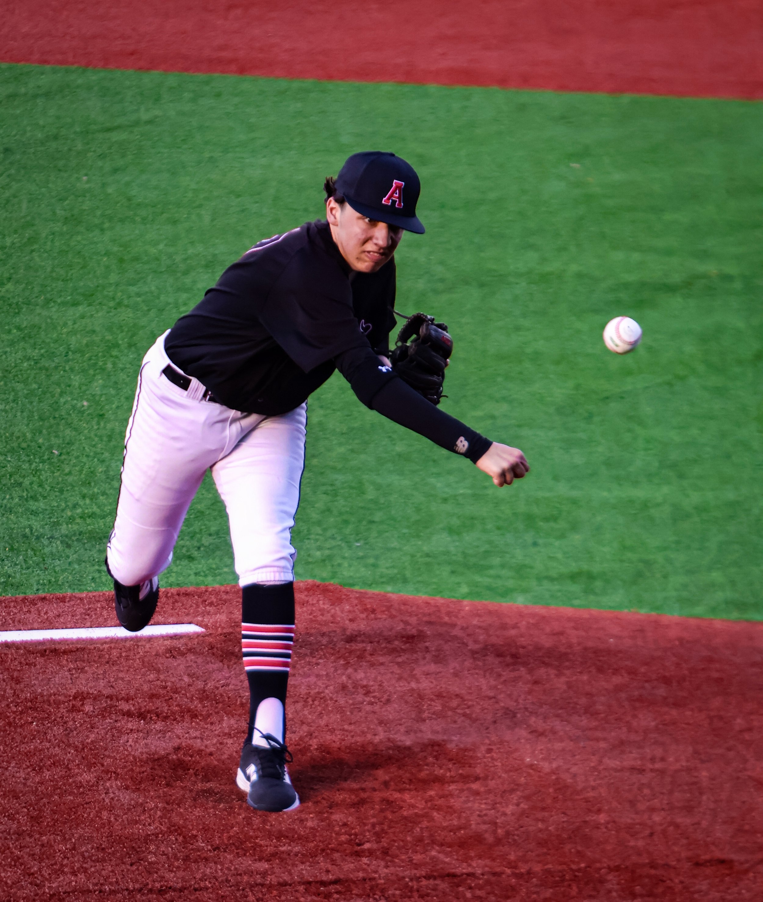 A baseball player in a black uniform and cap with a red 'A' logo, batting on a baseball field with green grass and red dirt, as the ball approaches.