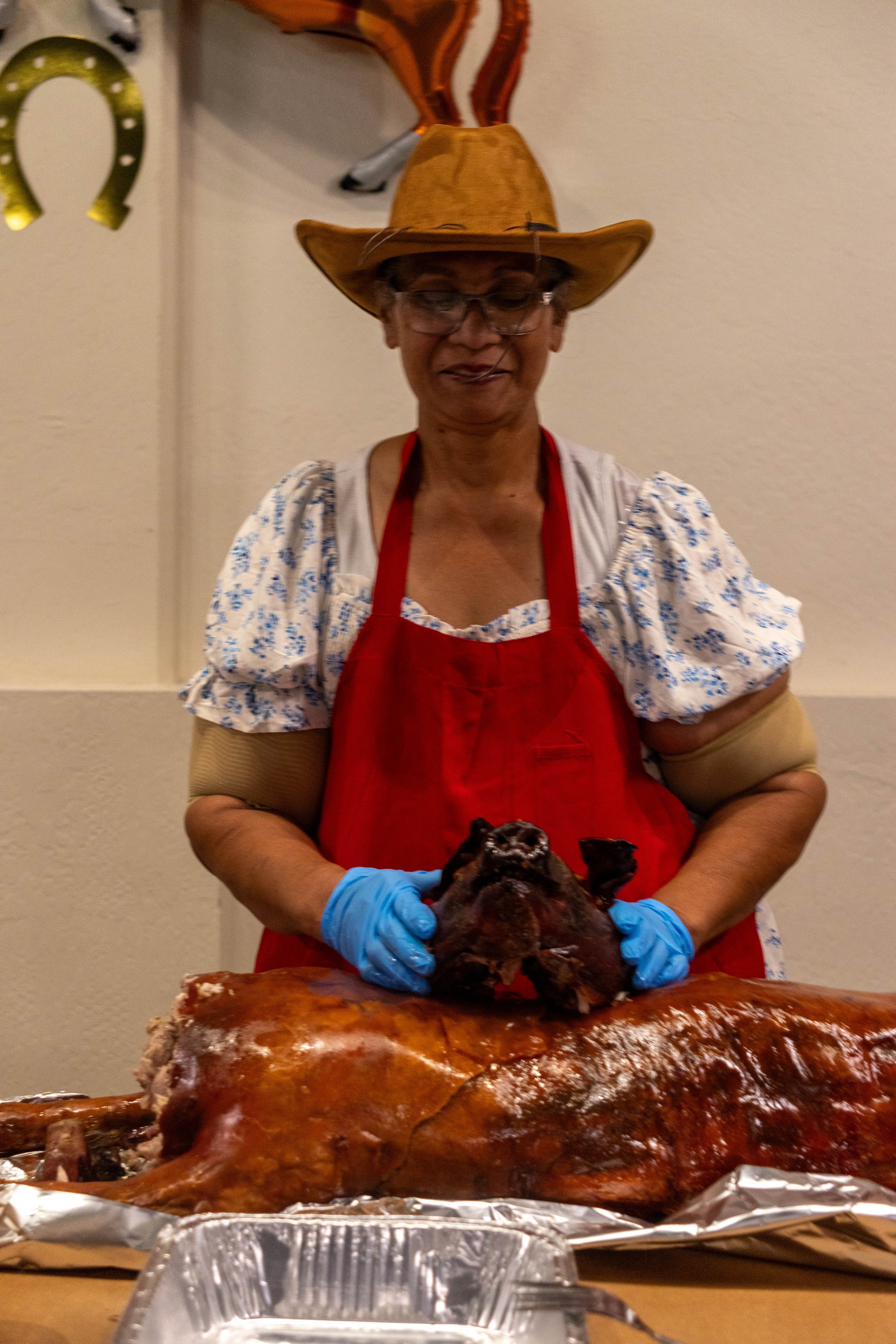 A woman wearing glasses, a straw hat, a red apron, and blue gloves, is holding a roasted pig's head in front of a large glazed ham on a table.