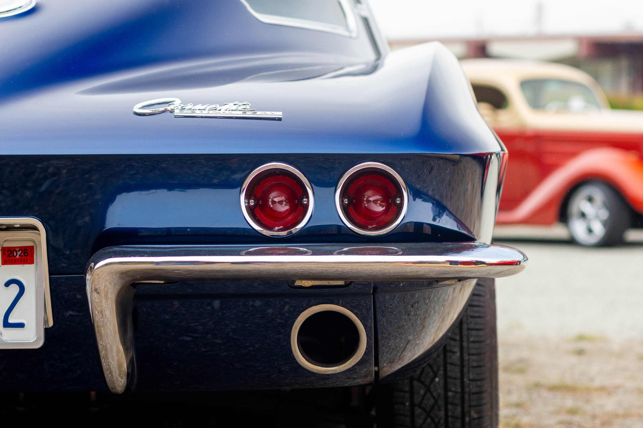 Close-up of the rear of a vintage blue Chevrolet car showcasing tail lights, chrome bumper, and exhaust pipe with other classic cars in the background.