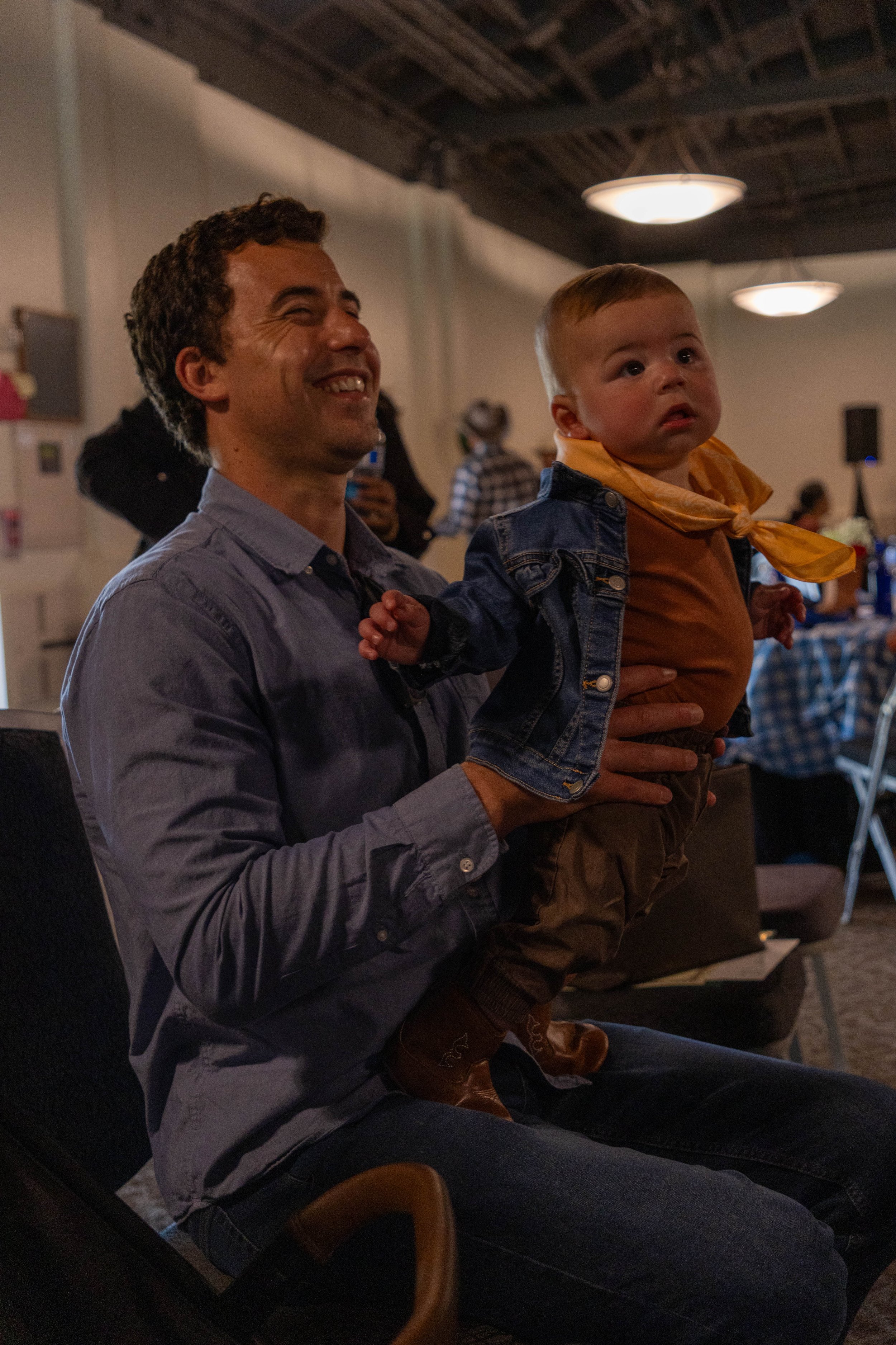 A man is holding a young child on his lap at an indoor gathering or event. The man is smiling and the child appears to be looking curiously at something. The setting includes other people in the background, and the lighting is warm and ambient.