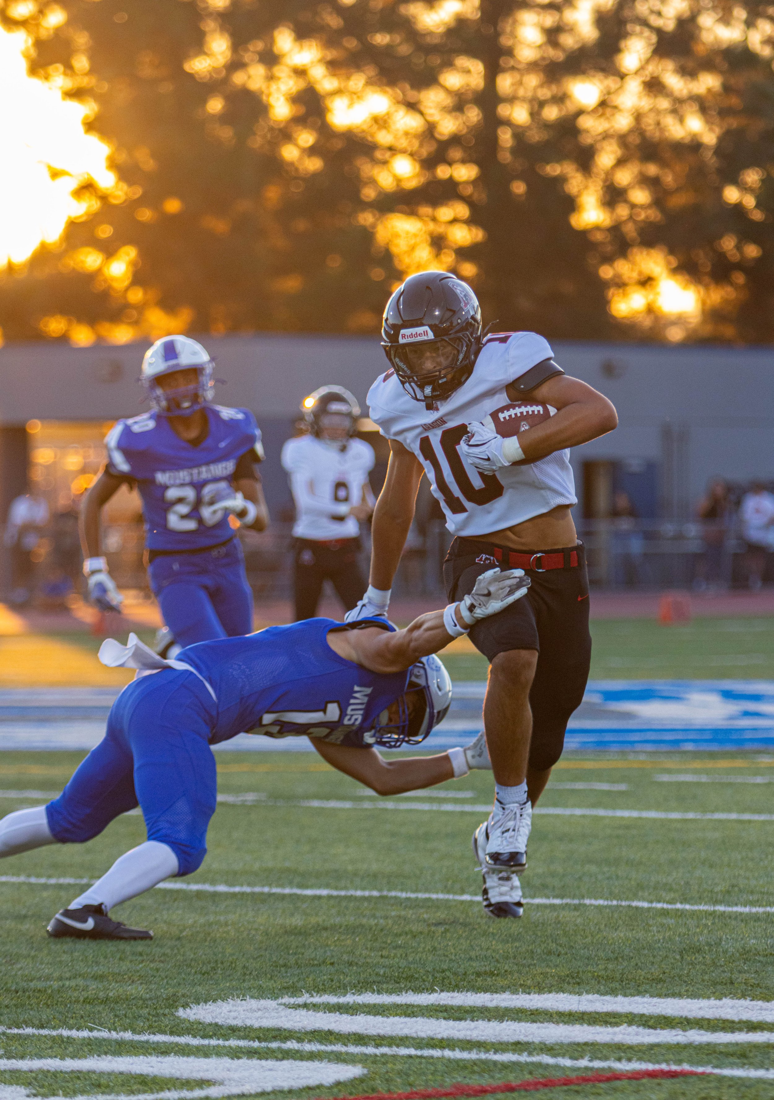 A football game at sunset with players in action, one in black and white uniform carrying the ball, a player in blue attempting to tackle him, and another player in white and blue in the background.