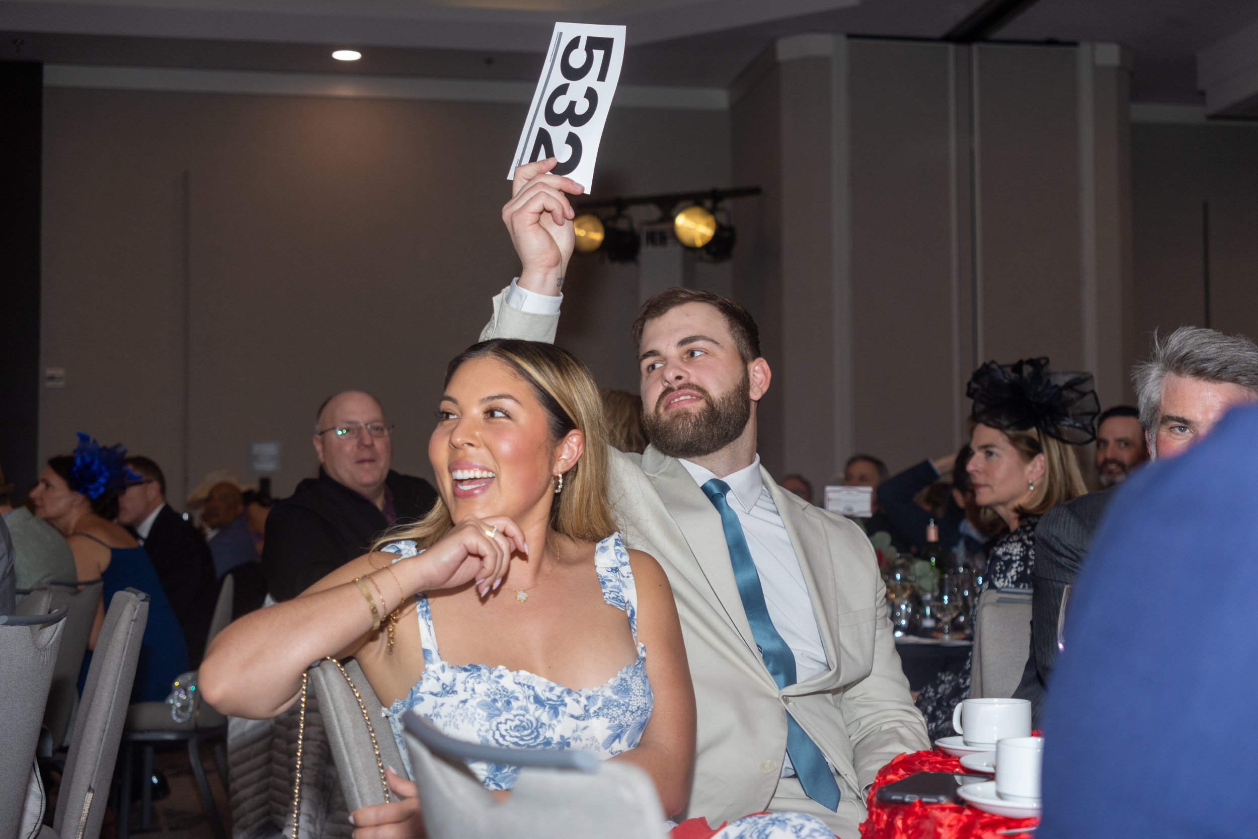 A man and a woman sitting at a formal event, the woman smiling and the man raising an auction paddle with the number 532.
