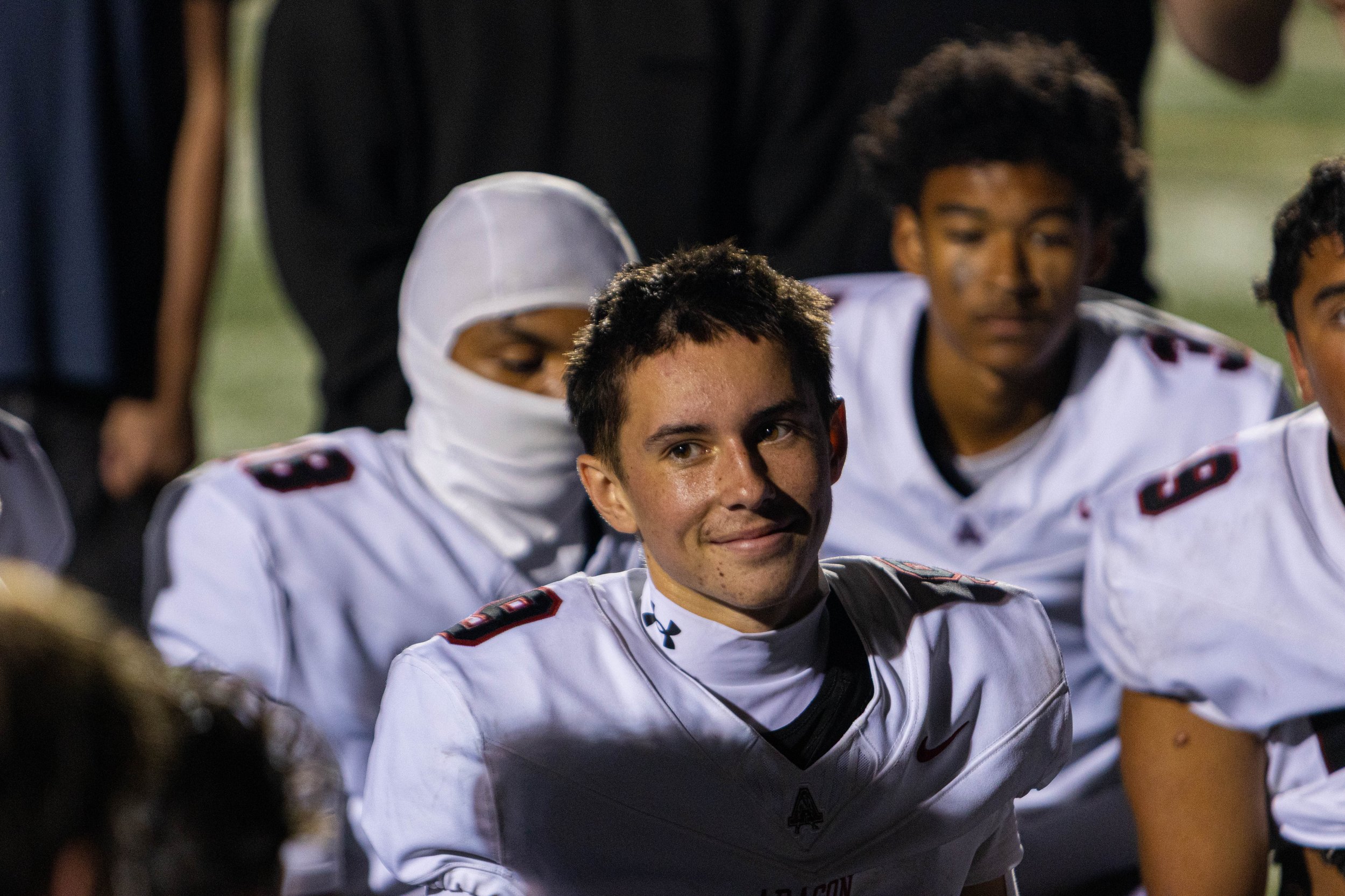 Group of young football players, some wearing white jerseys with red and black accents, gathered together during a game or practice.