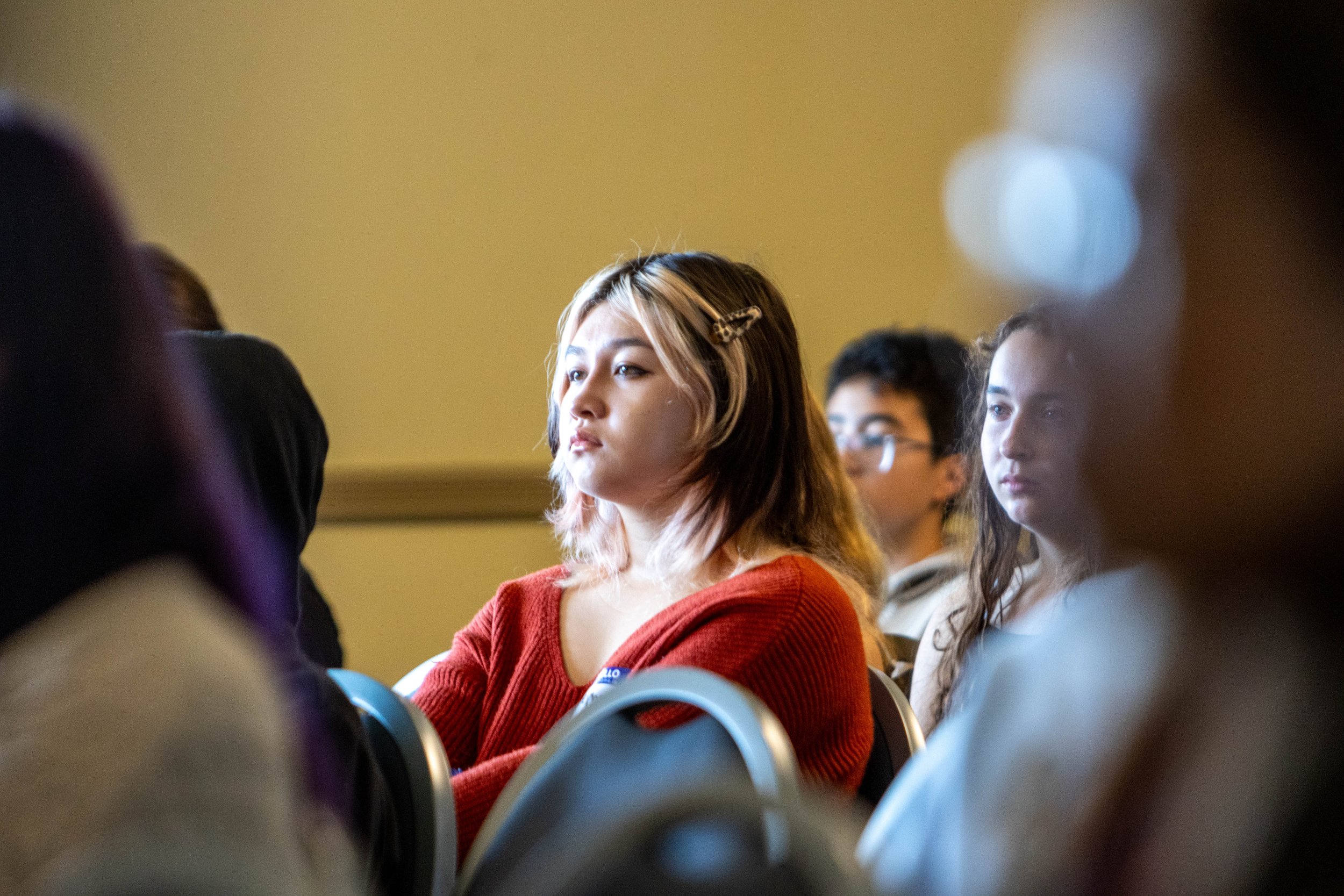 A young woman with blonde hair and a hair clip, wearing a red sweater, sitting in a crowded room with people sitting in chairs behind her.