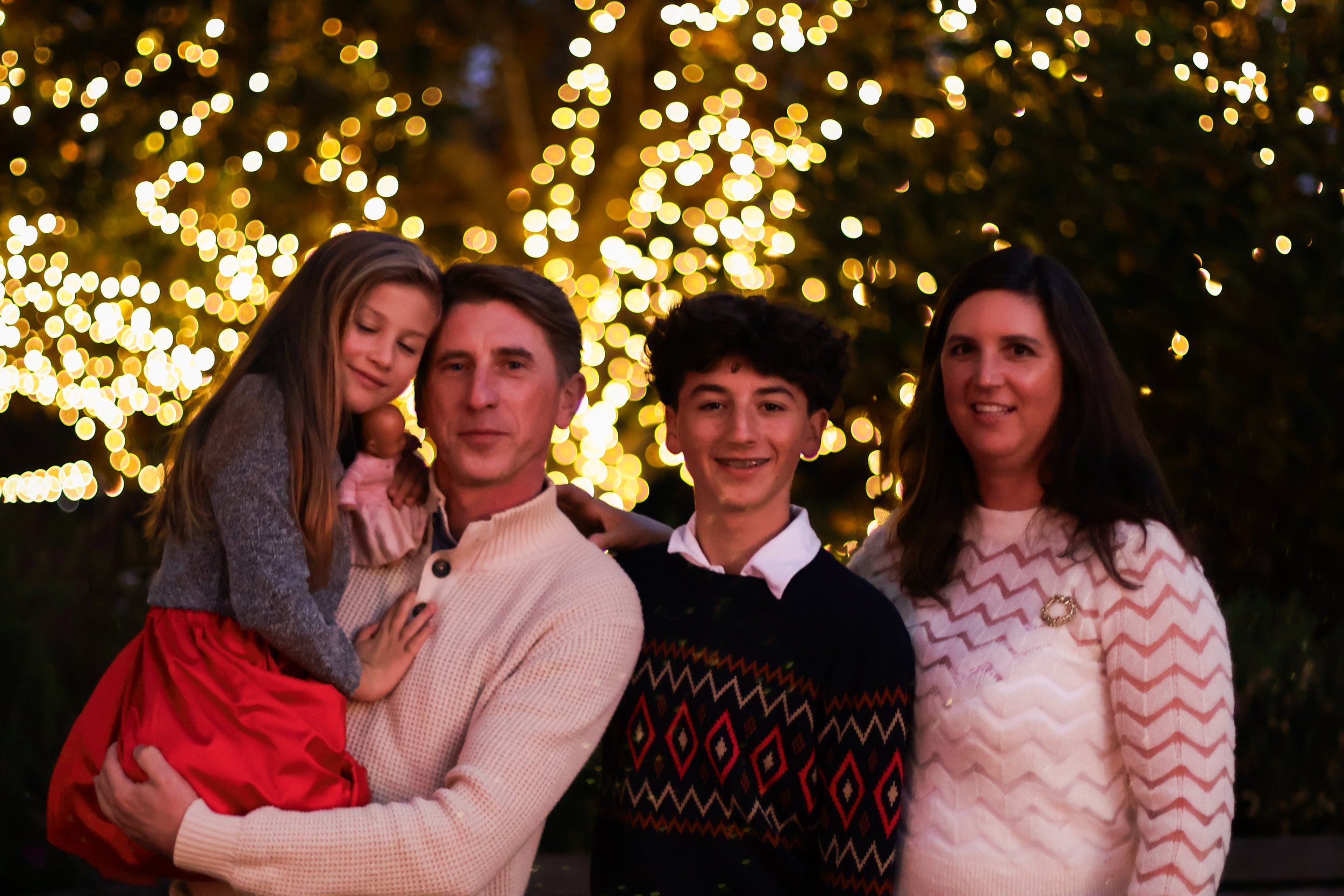Family of four posing outdoors at night in front of a decorated Christmas tree with yellow lights, with a man, woman, teenage boy, and young girl.