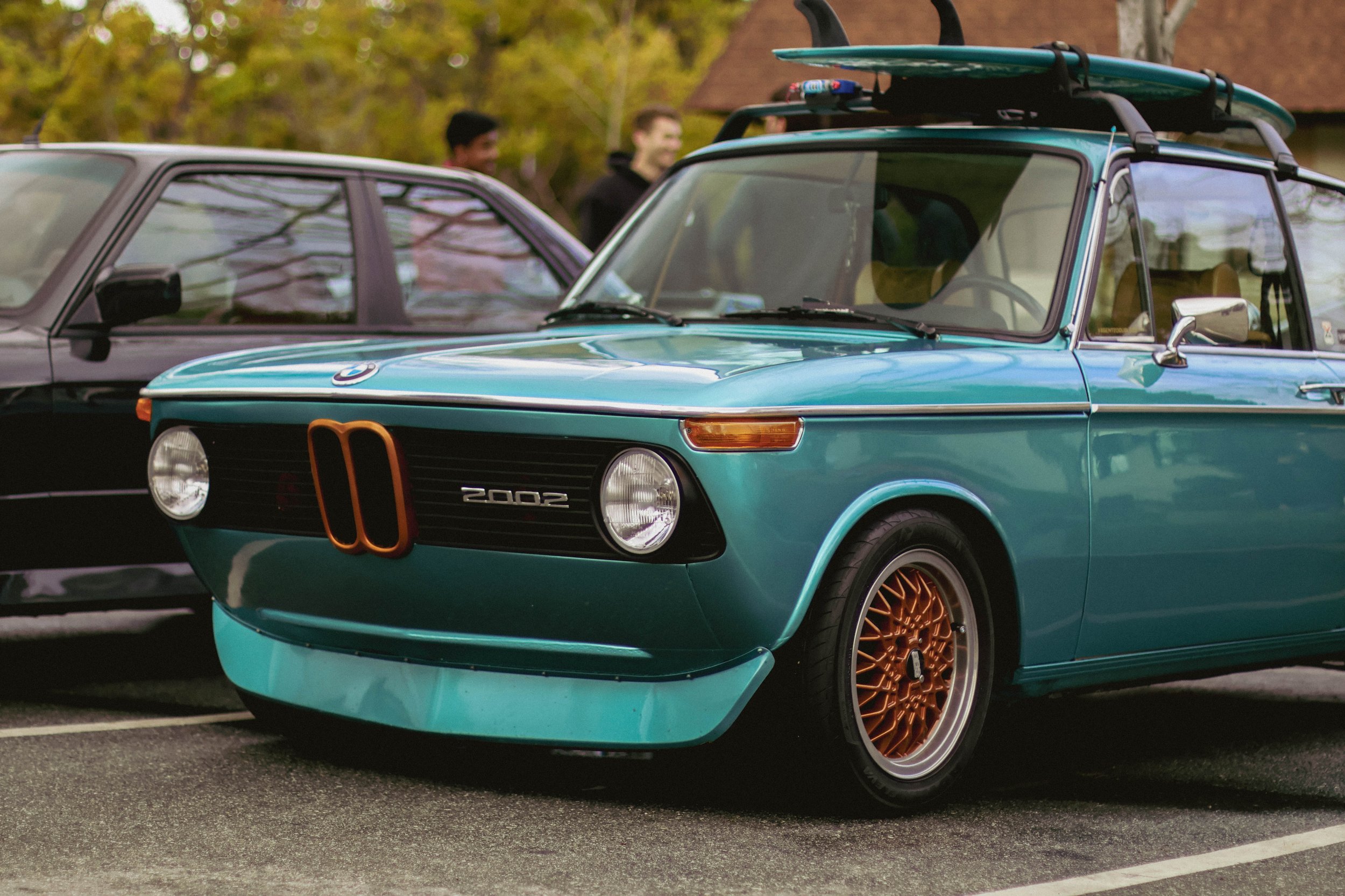 A teal vintage BMW 2002 car with a surfboard on the roof parked in a lot, with people and blurred trees in the background.