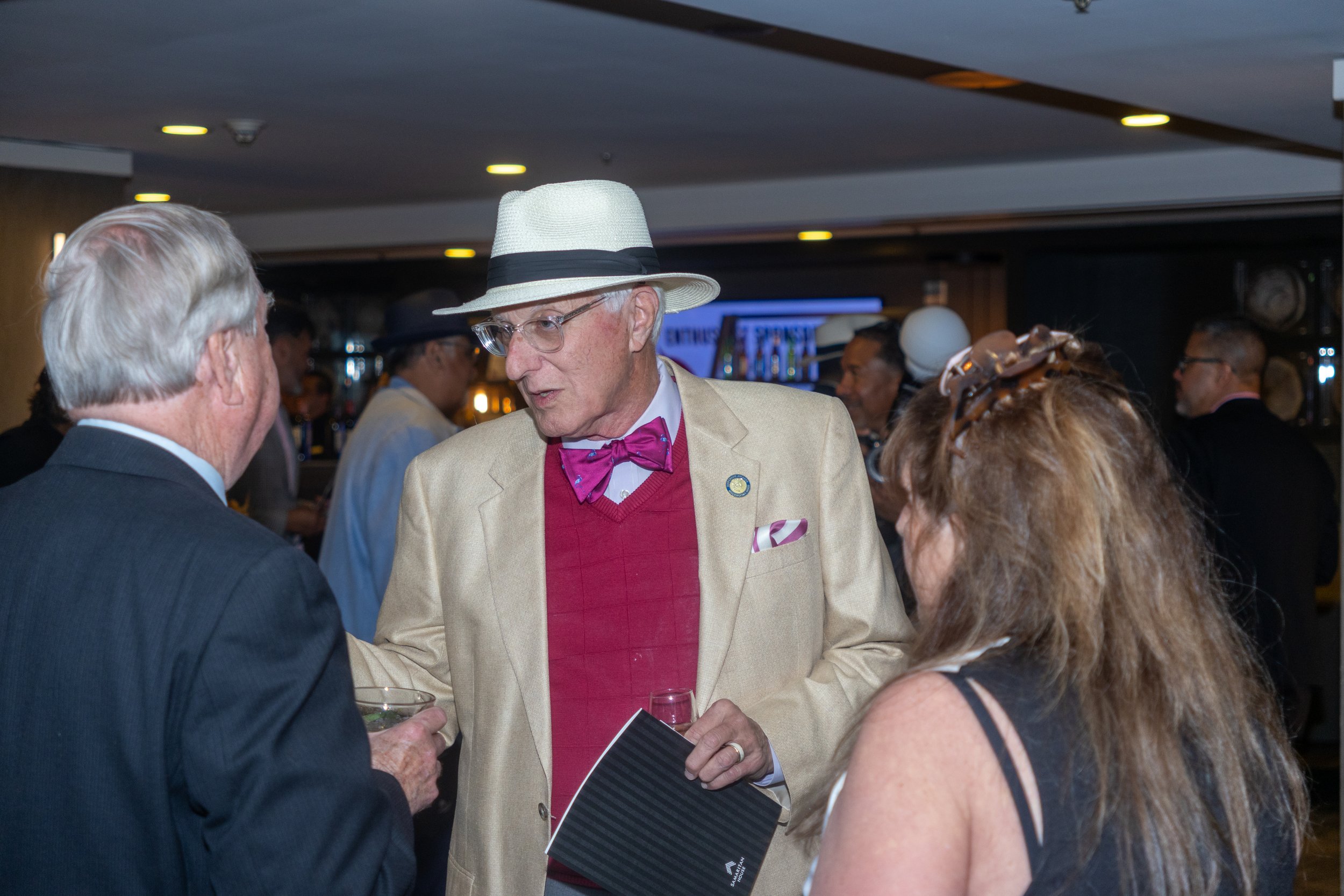 A man in a beige blazer, red sweater, pink bow tie, and a white hat with a black band is engaged in conversation in a crowded indoor event, holding a black folder in one hand. Other people are visible in the background.