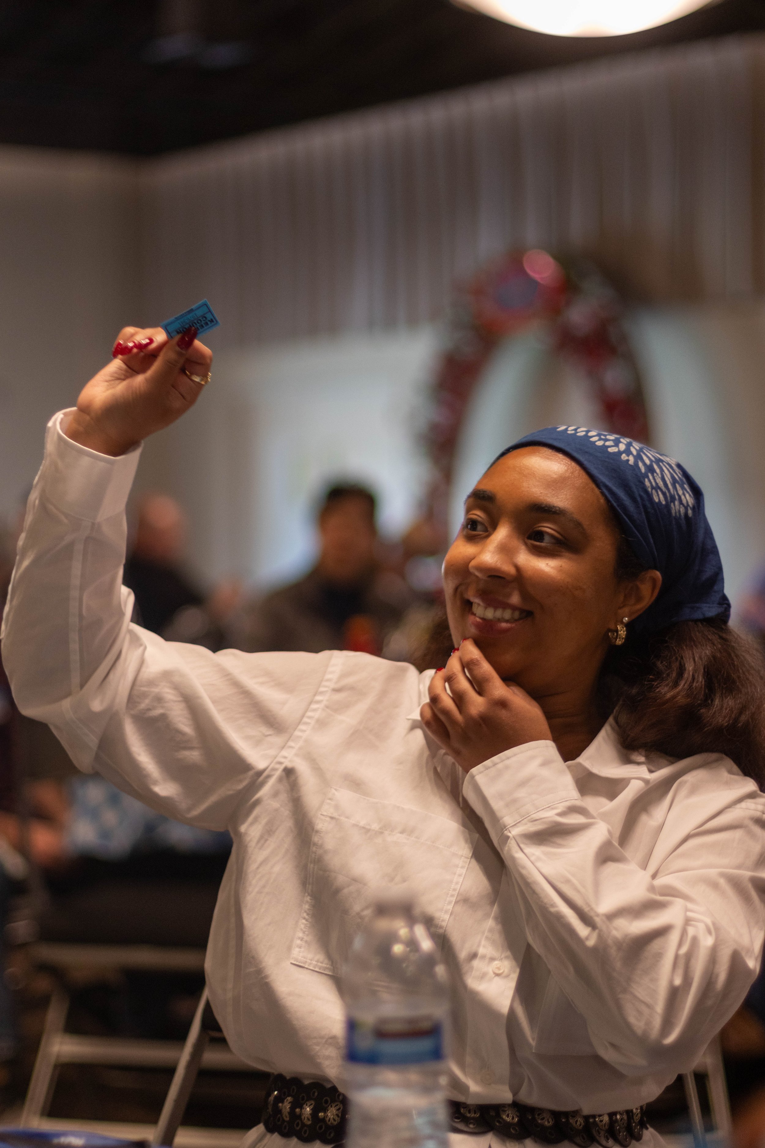 A woman smiling and taking a selfie with her phone. She is wearing a white shirt and a blue headscarf, with a festive holiday wreath in the background.