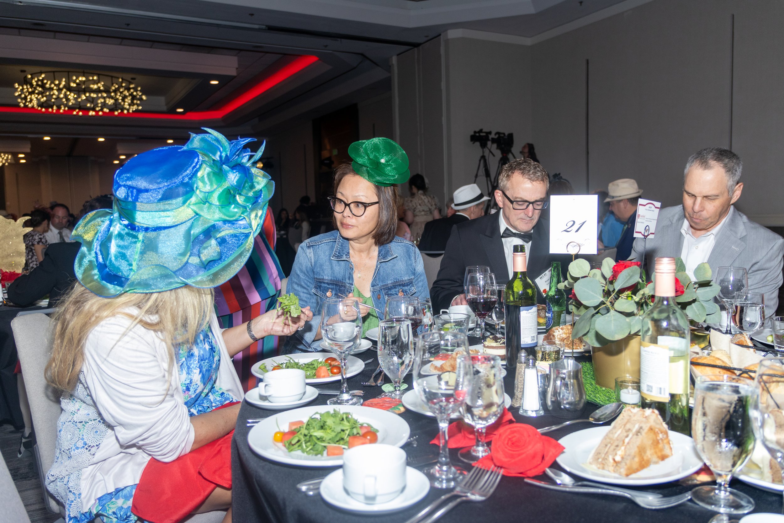 People sitting at a banquet table during the 2026 Samaritan House Gala decorated with flowers, dressed in formal attire, some wearing large colorful hats, in a decorated banquet hall with dim lighting.