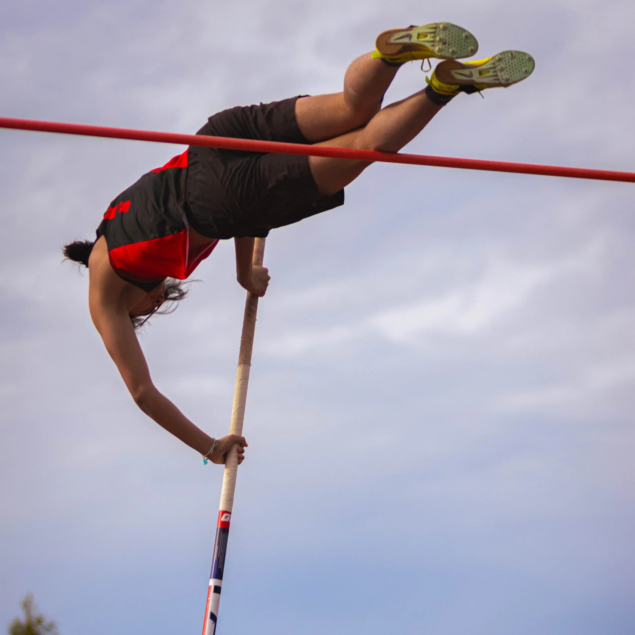 A pole vaulter in mid-air clearing the bar during a track and field event, with a cloudy sky in the background.