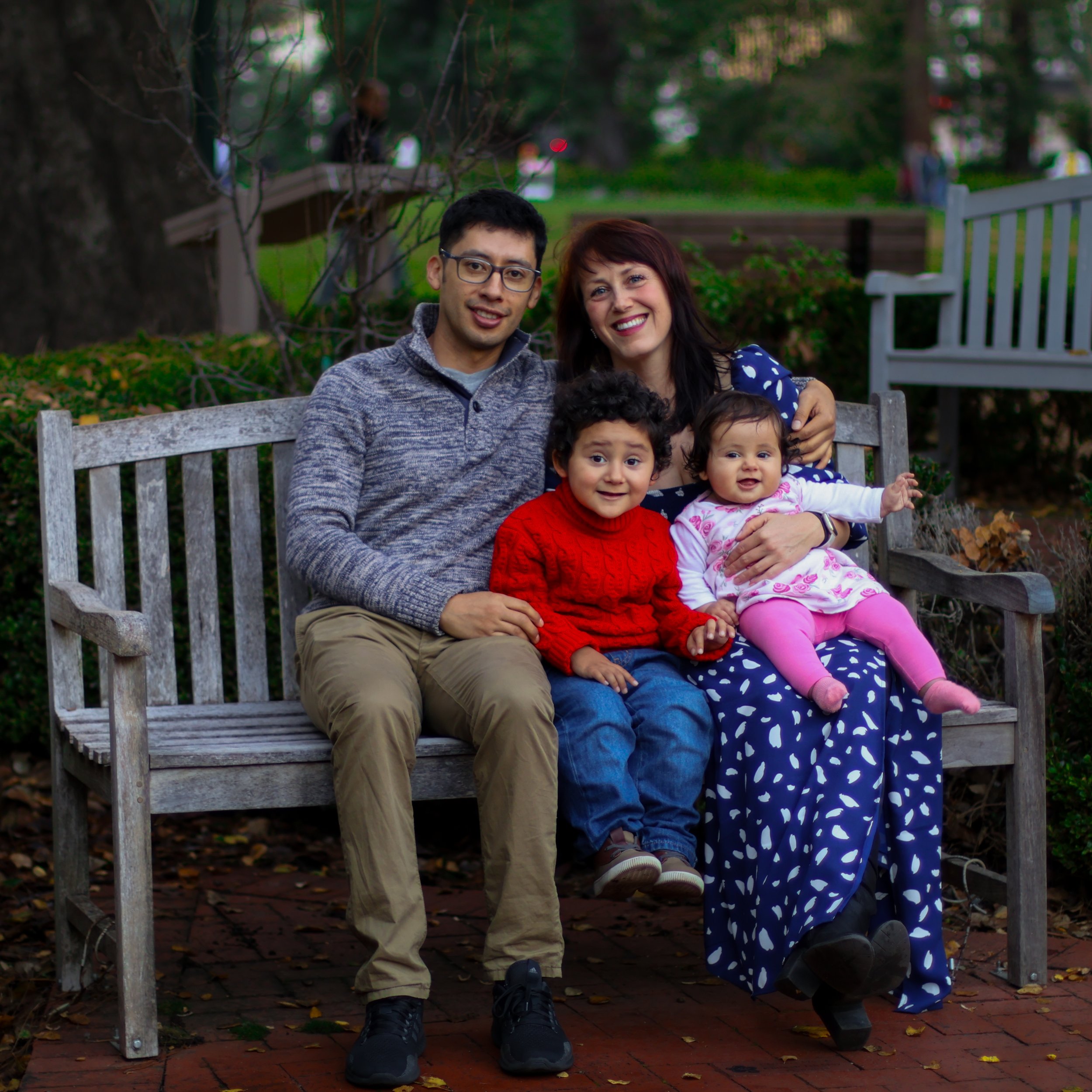 A happy multi-generational family sitting on a park bench, smiling at the camera with trees and fallen leaves in the background.