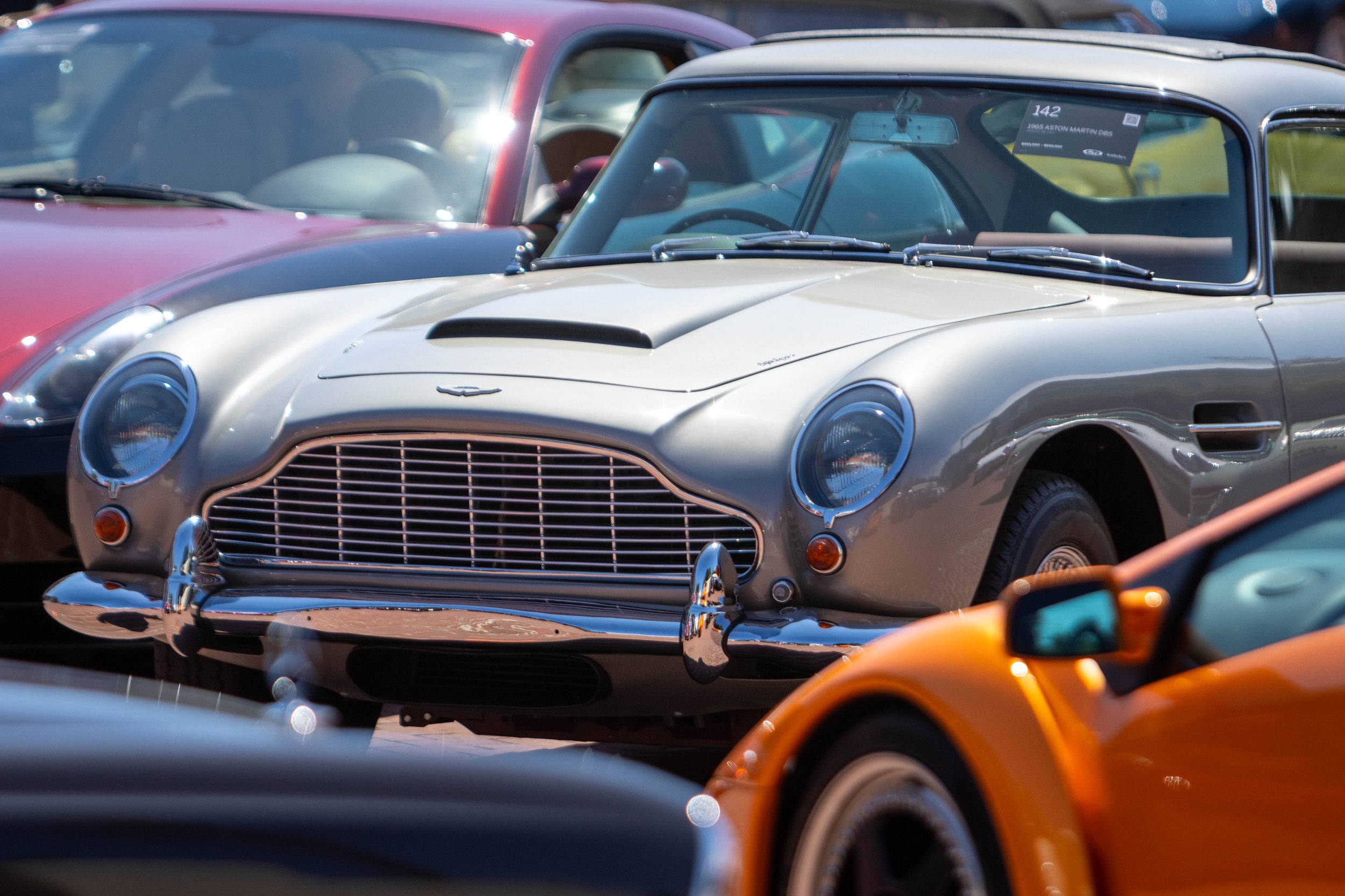 A vintage silver Aston Martin DB5 car parked among other cars, with a photo ticket visible on the windshield, in a parking lot.