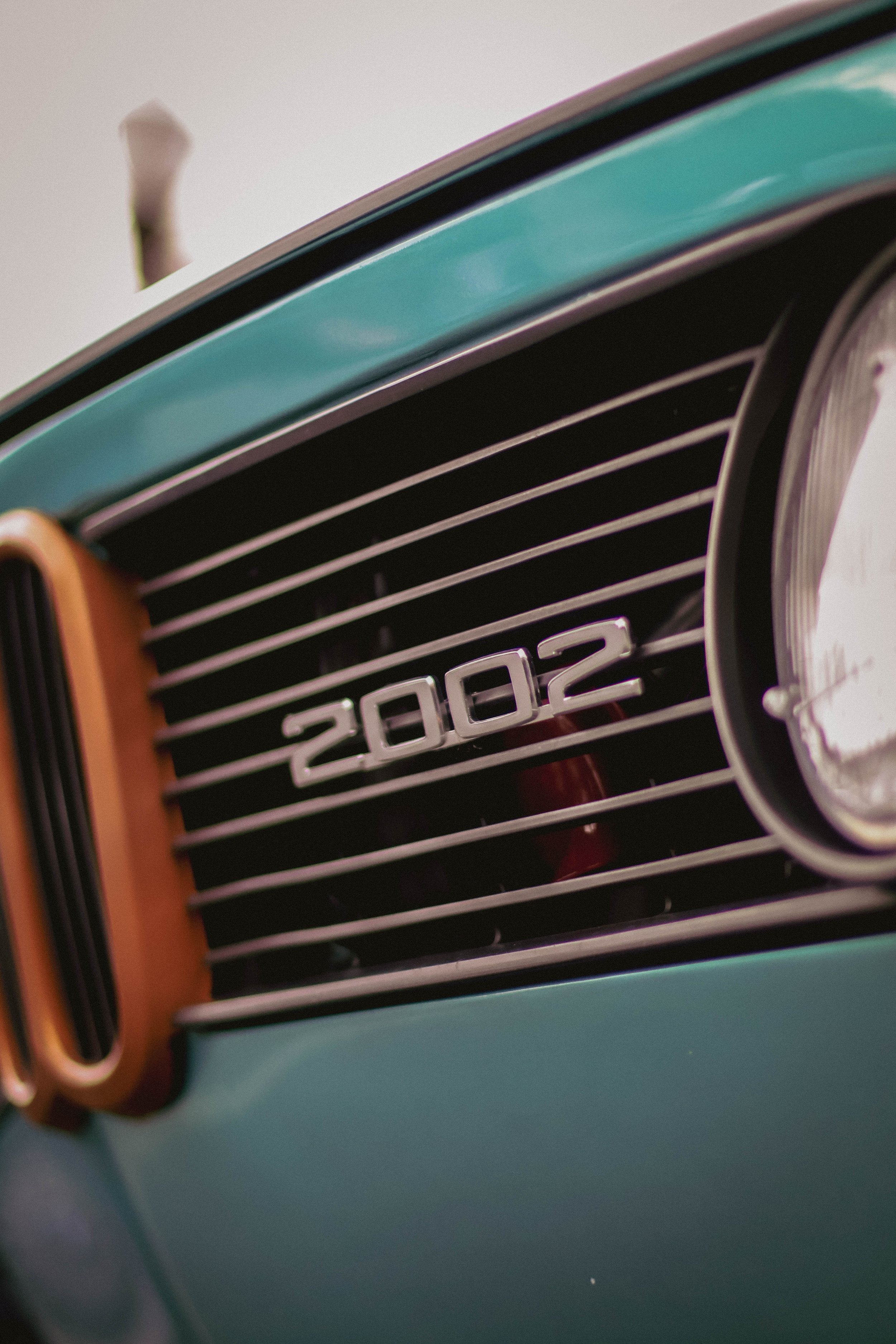 Close-up of a vintage teal-colored vehicle's front grille with horizontal black slats and the number "2002" visible, partially showing the round headlight on the right side.