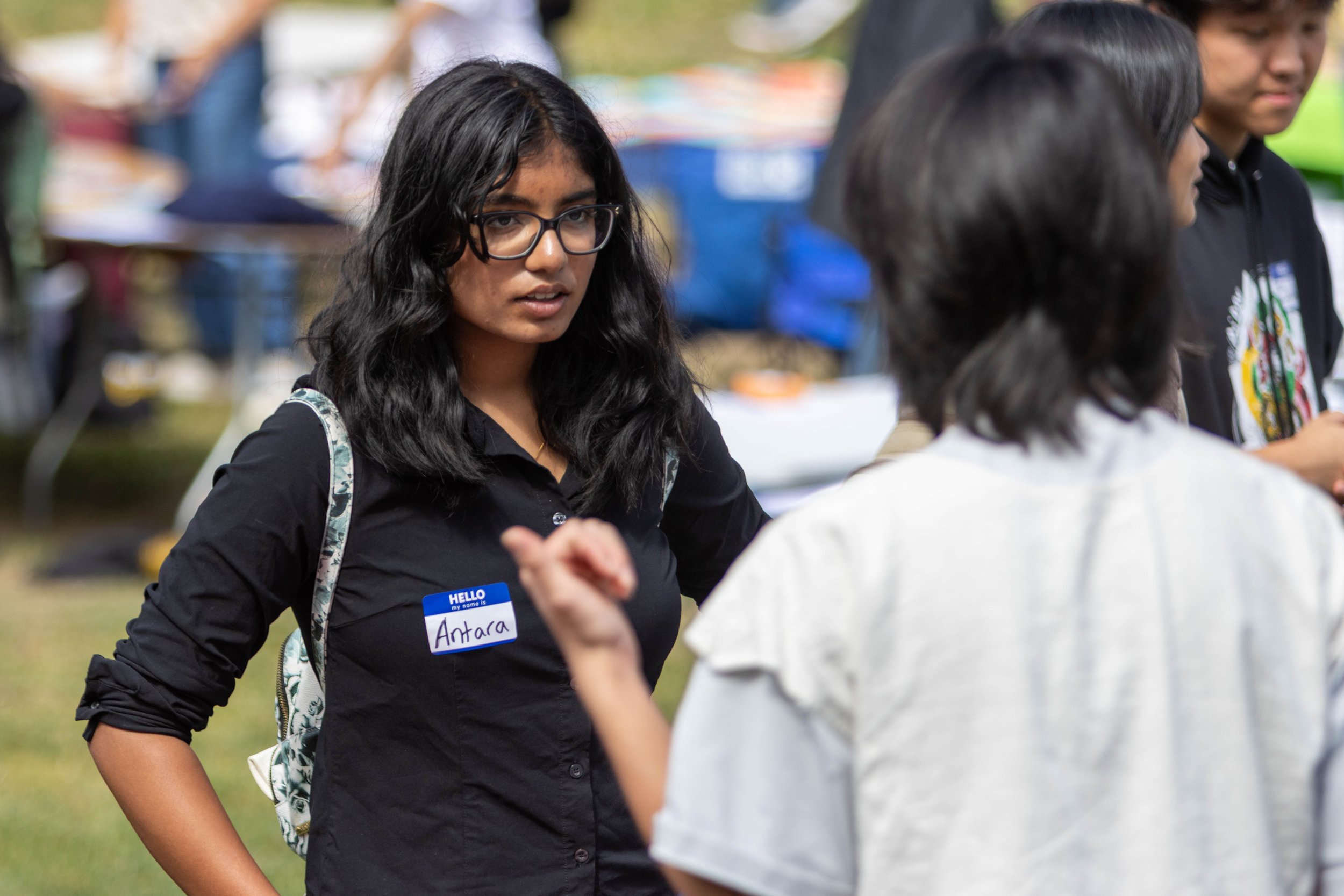 A young woman with glasses and long dark hair, wearing a black shirt and a name tag that says 'Antara', participating in an outdoor event; she is looking at a person in front of her.