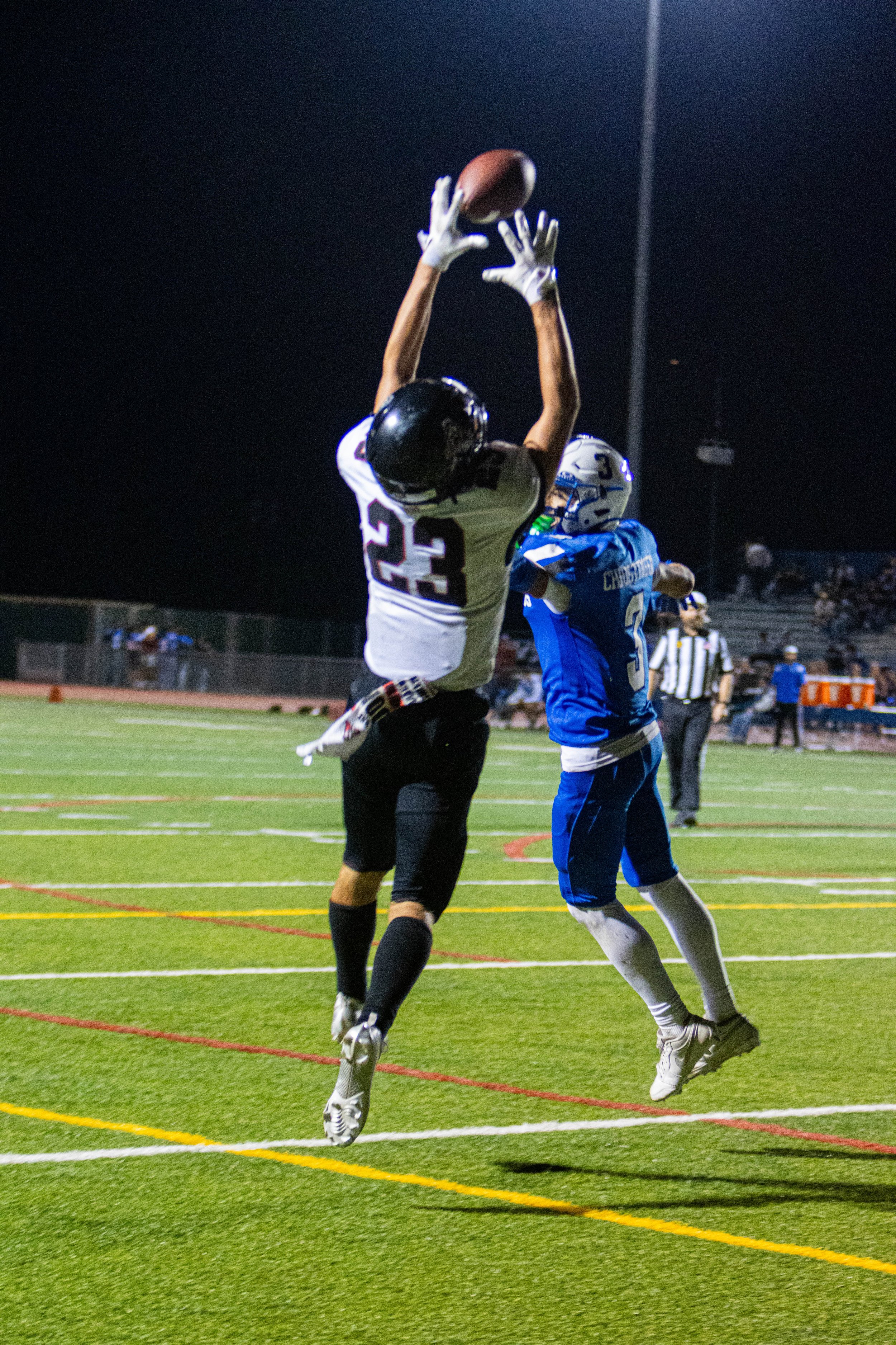 A nighttime football game showing two players jumping to catch the ball, surrounded by a football field with yard lines, a referee, and spectators in the background.