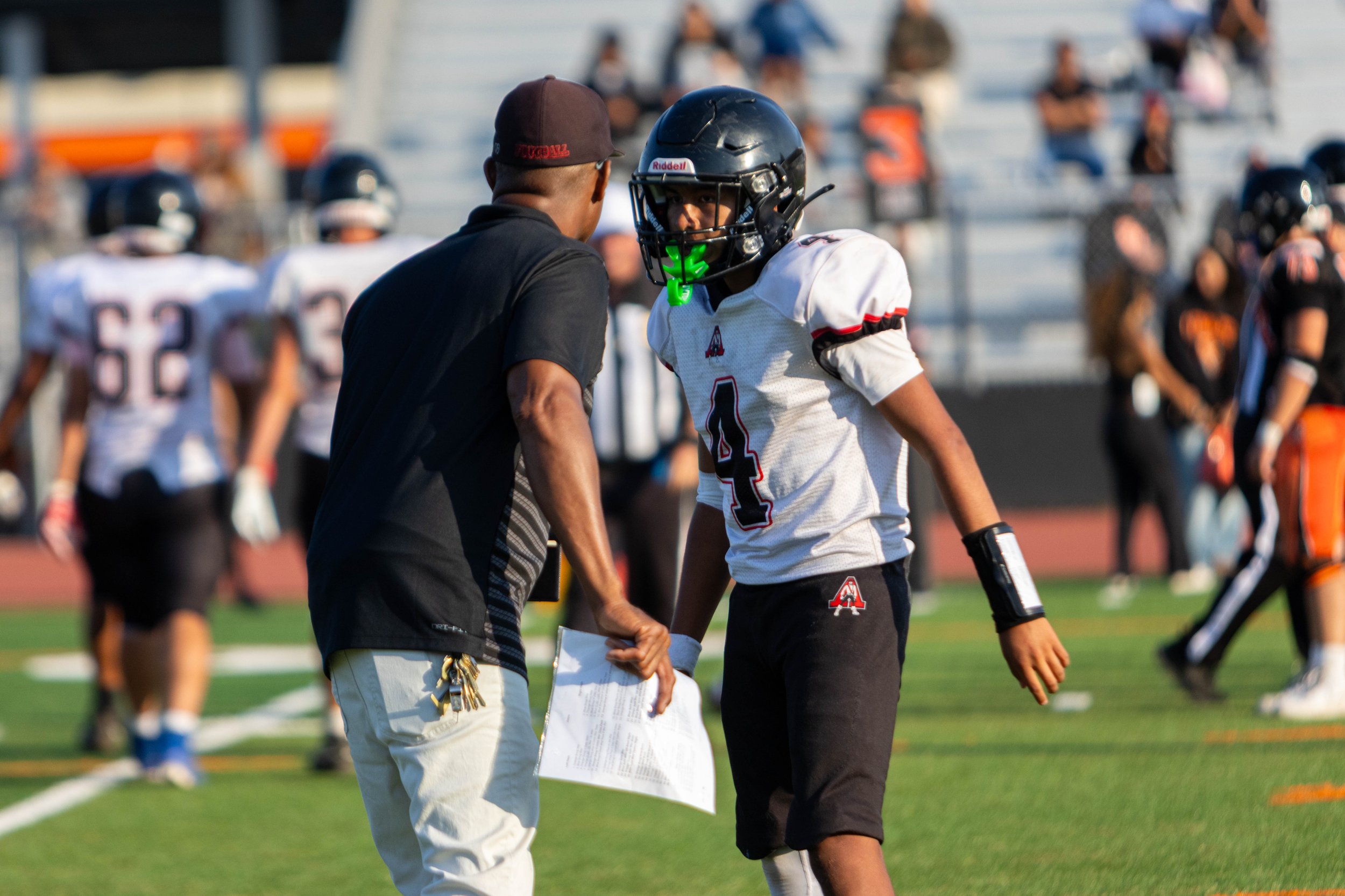 Football coach talking to player on the sidelines during a game on a sunny day.