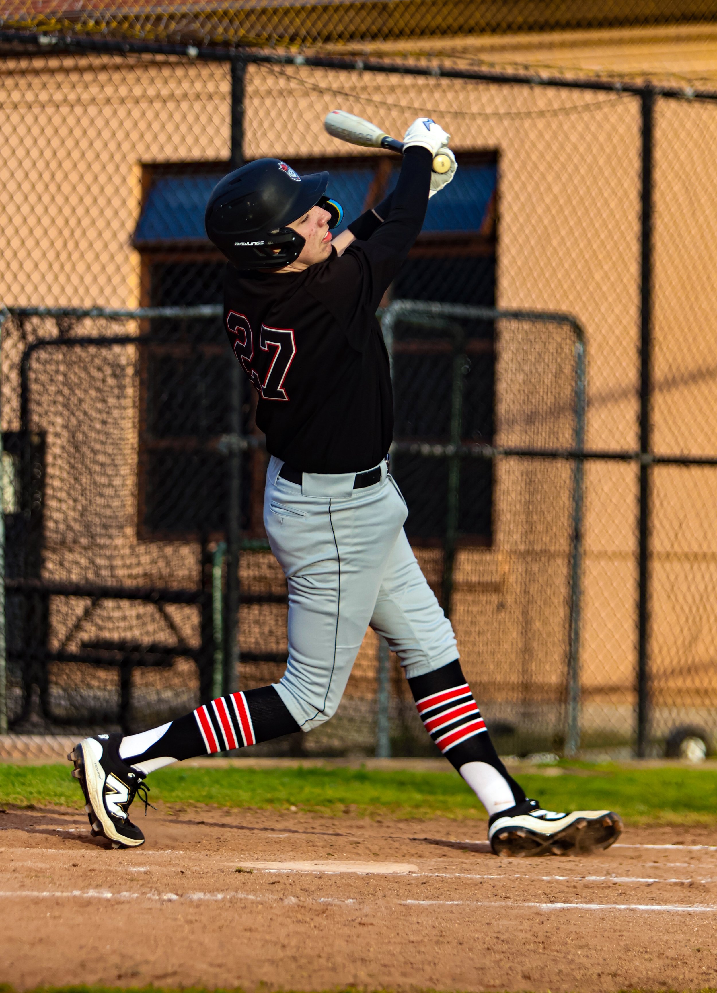 A baseball player in black uniform with number 27, wearing a helmet and striped socks, is running towards first base on a baseball field, with a chain-link fence and a building in the background, during sunset.
