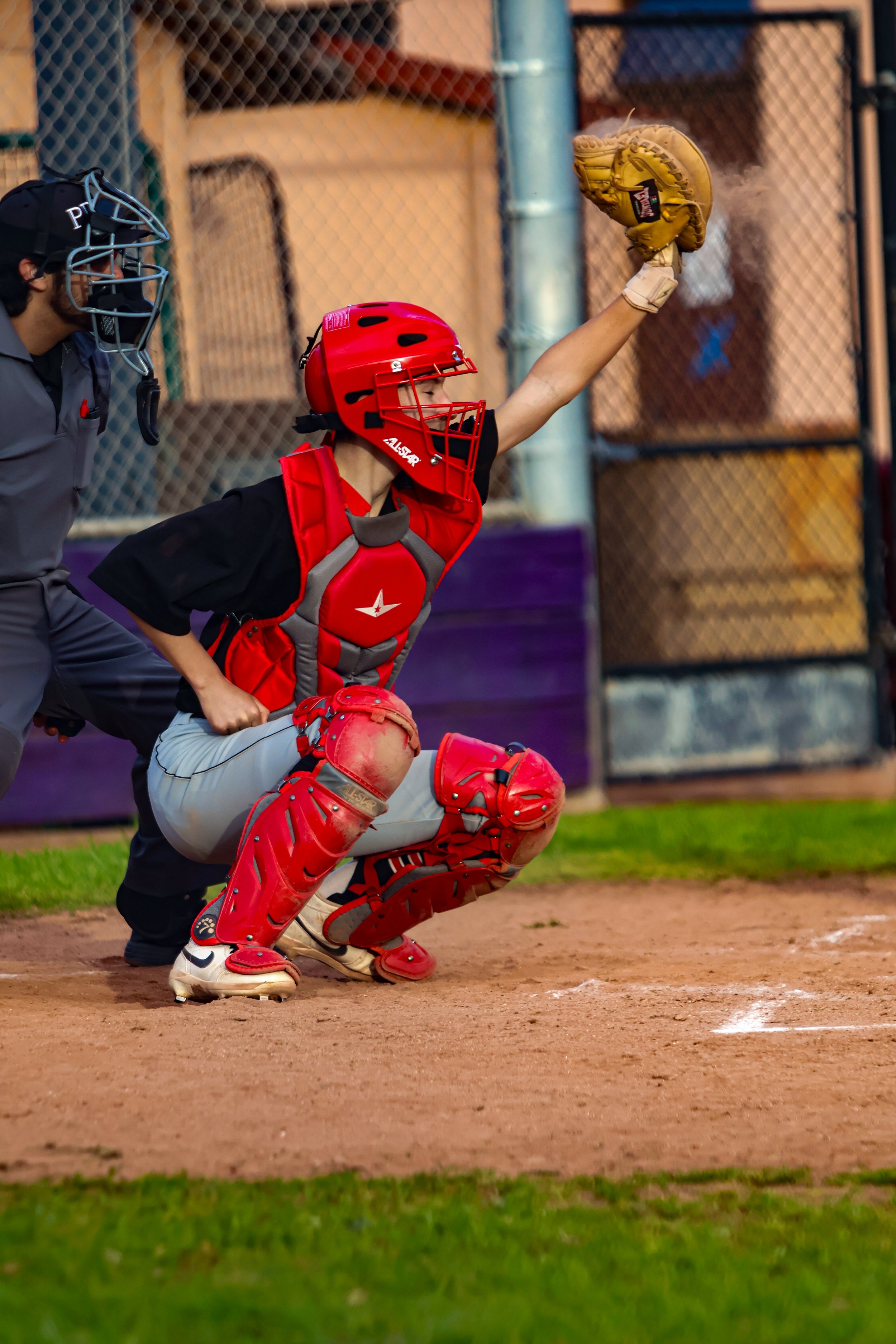 A youth baseball catcher wearing red gear, a helmet, and a glove, is crouching behind home plate while a coach or umpire stands nearby. The catcher is reaching up with his glove to catch a ball, with a baseball field and chain link fence in the backg