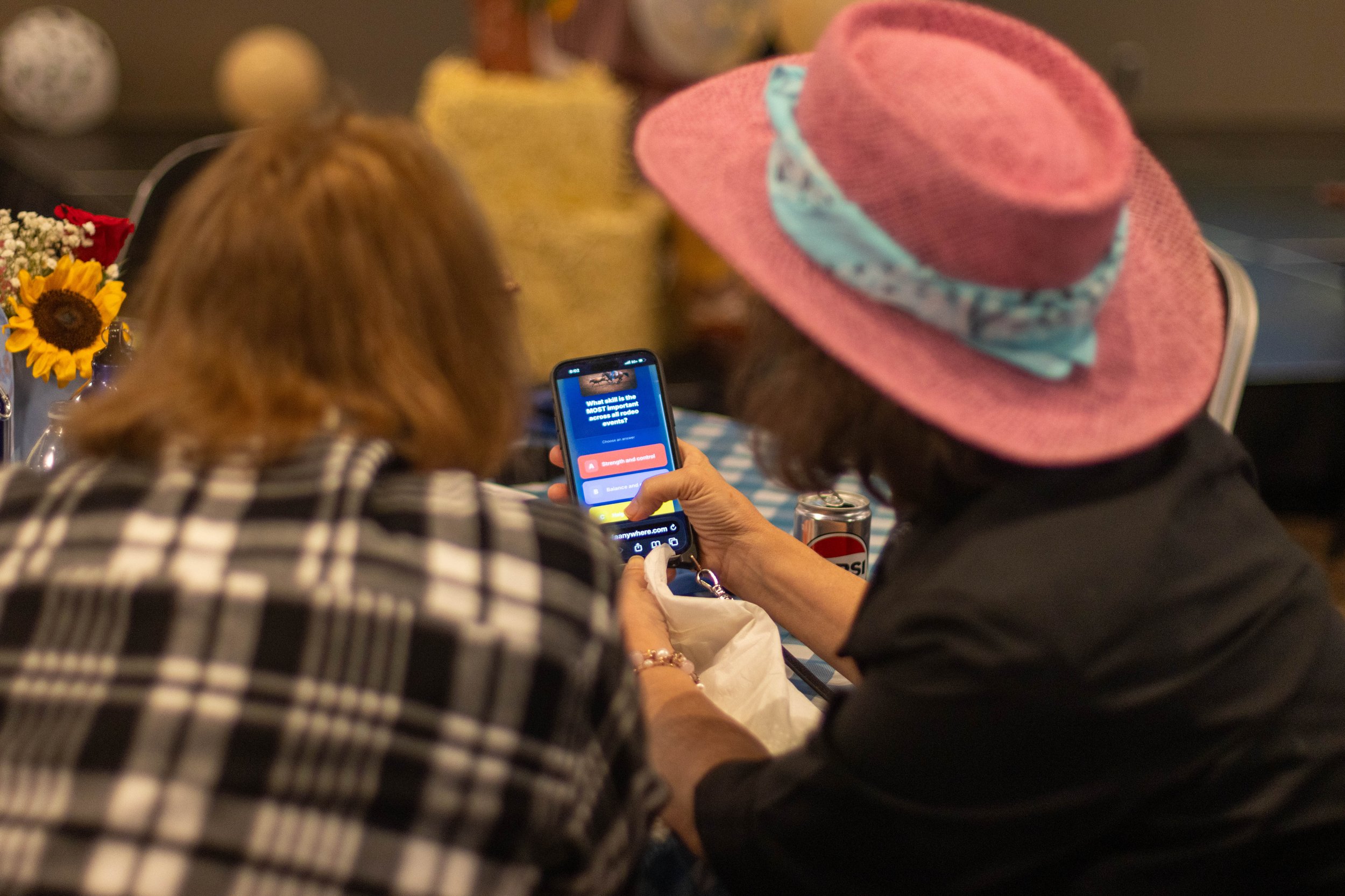 A group of people sitting at a table, with a woman in a pink hat in the foreground, looking at her phone. The table has a sunflower and other flowers, and the background appears to be a casual indoor setting.