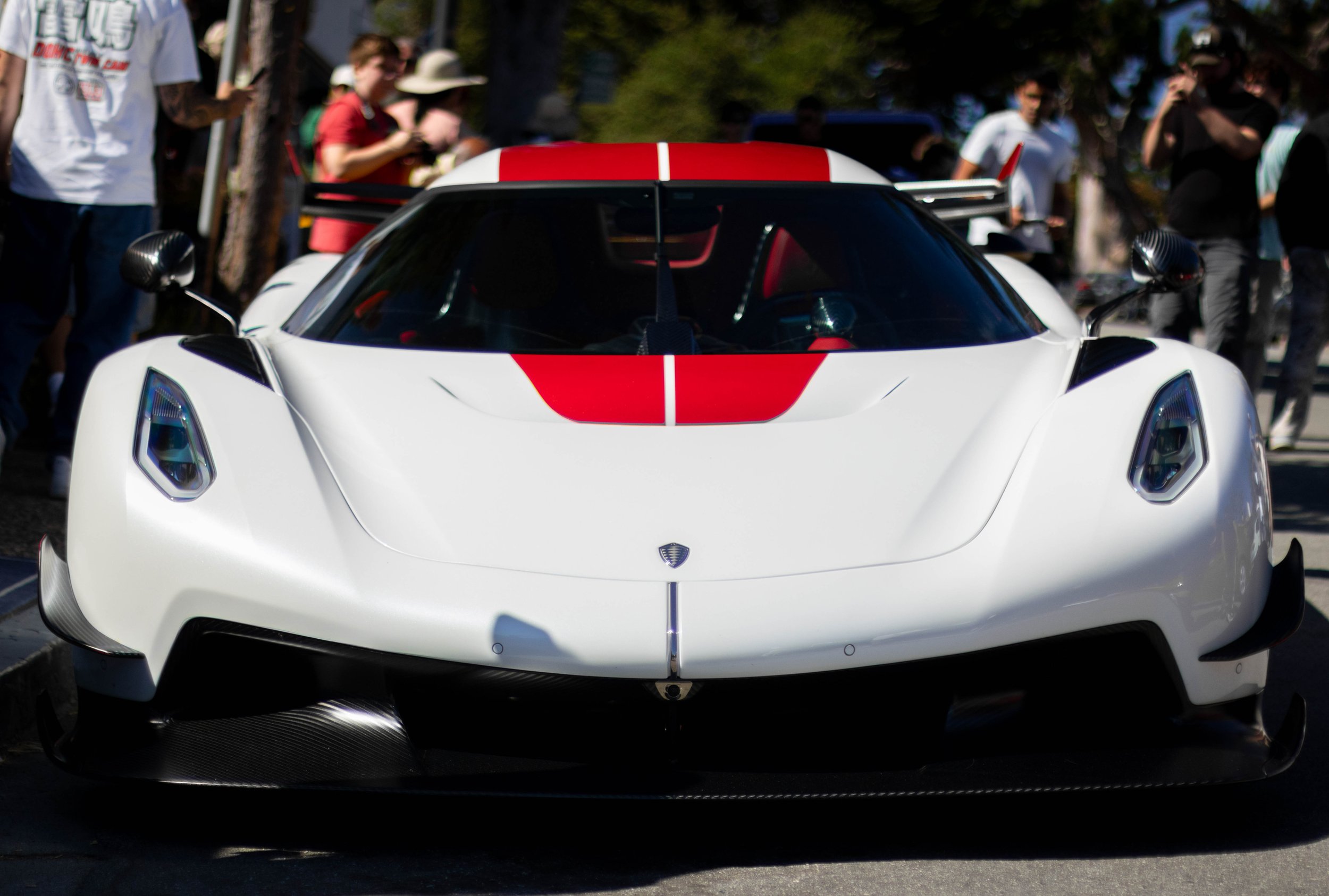 A white sports car with red accents on the hood and roof, surrounded by people at an outdoor car event.