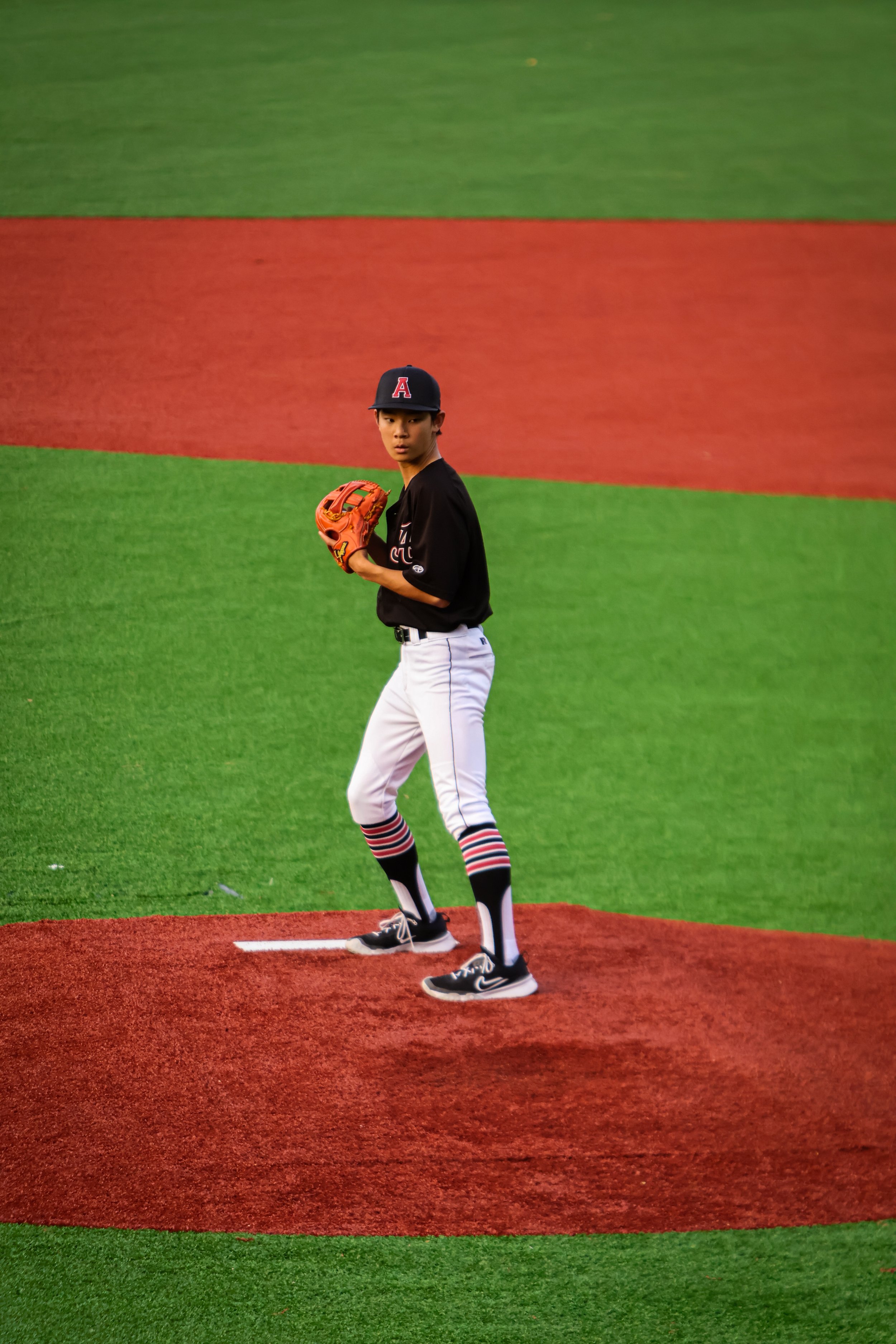 A baseball player standing on the pitcher's mound, holding a glove, wearing a black jersey with the letter A, white pants, and striped socks, looking towards the camera at a baseball field.