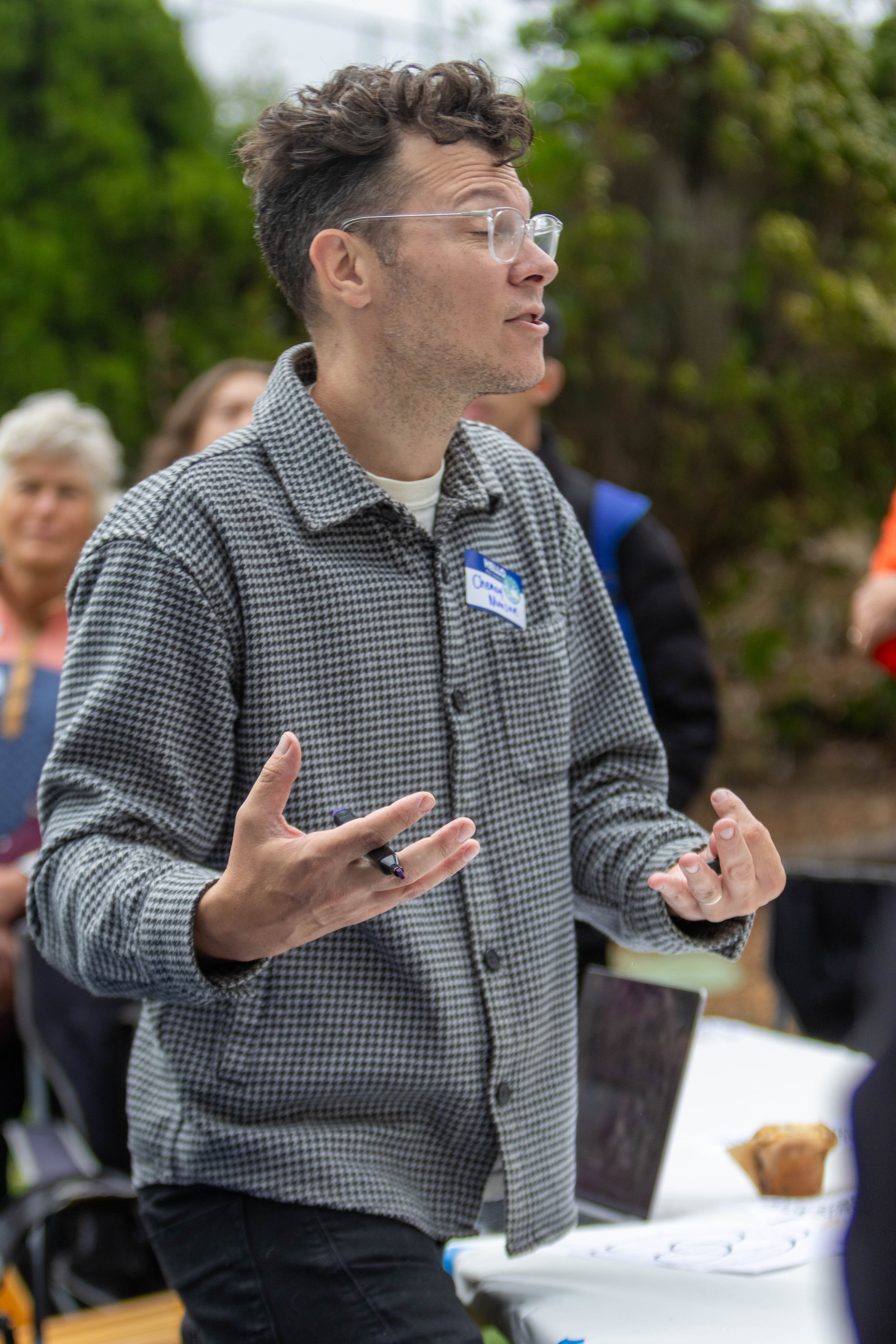 A young man with glasses, wearing a checkered shirt and a name tag, appears to be speaking or giving a presentation outdoors, with a blurred background of people and greenery.