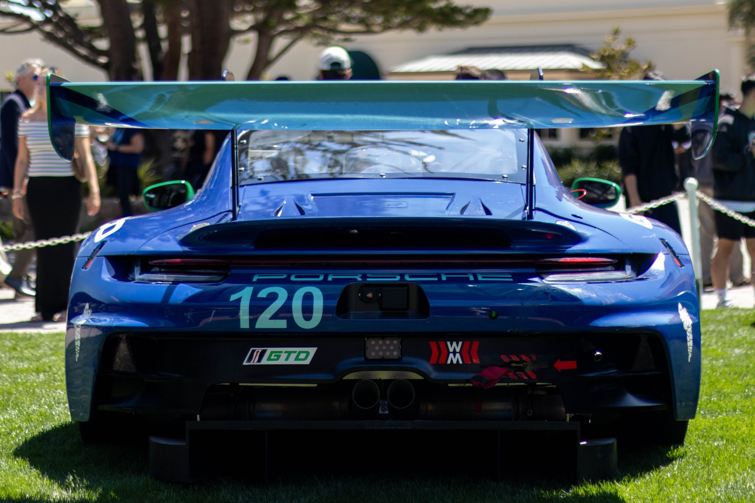 A blue race car with large rear wing on display outdoors, surrounded by people and onlookers.