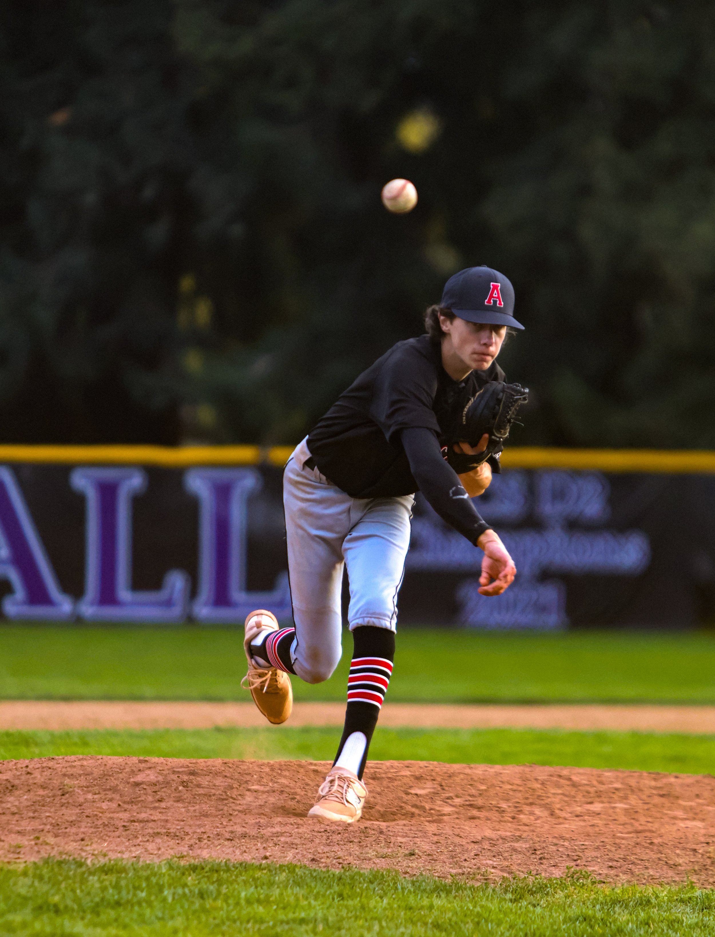 A young baseball player wearing a black jacket, gray baseball pants, striped knee-high socks, tan cleats, and a navy cap with a red letter 'A' is in the pitching position on a baseball field, preparing to throw the ball.