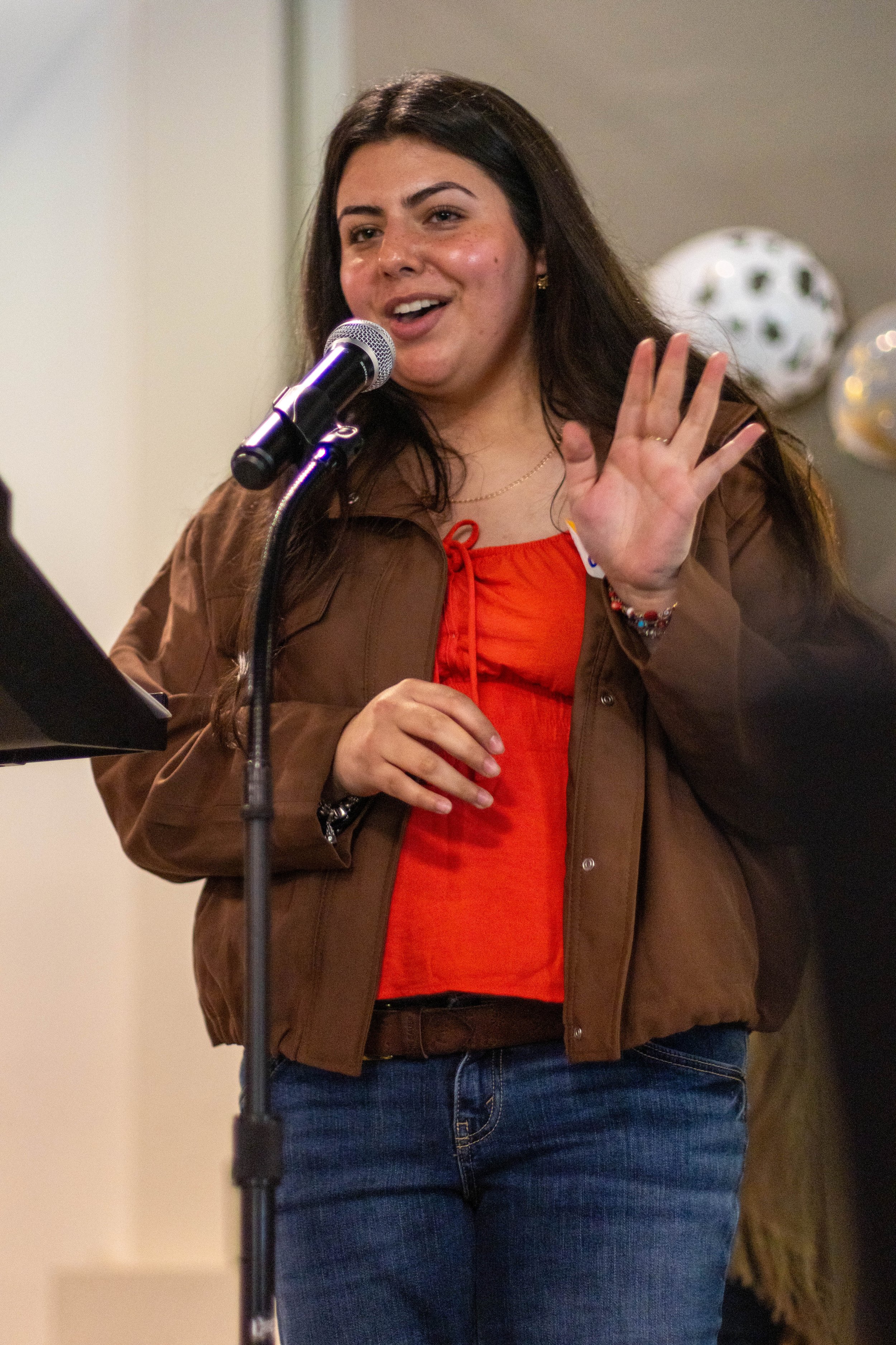 A woman with long dark hair speaking into a microphone, gesturing with her right hand, wearing a red top, brown jacket, and blue jeans, standing in front of a beige wall with decorative balloons in the background.