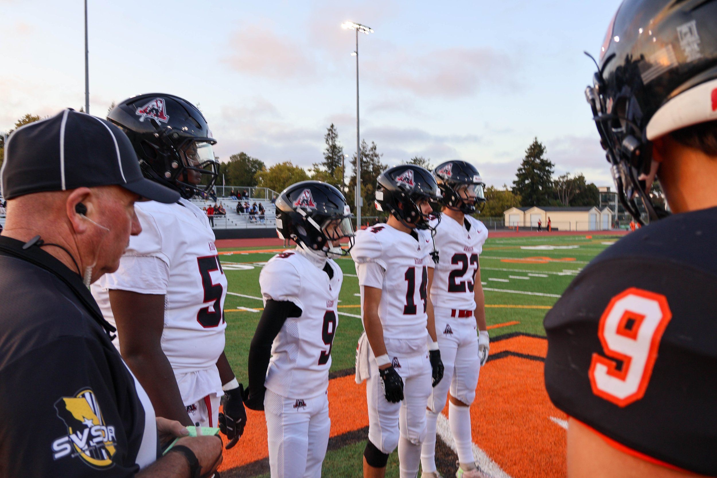 American football players and coaches standing in a huddle on a football field during a game or practice, wearing helmets and uniforms, with some on the sidelines and spectators in the background.