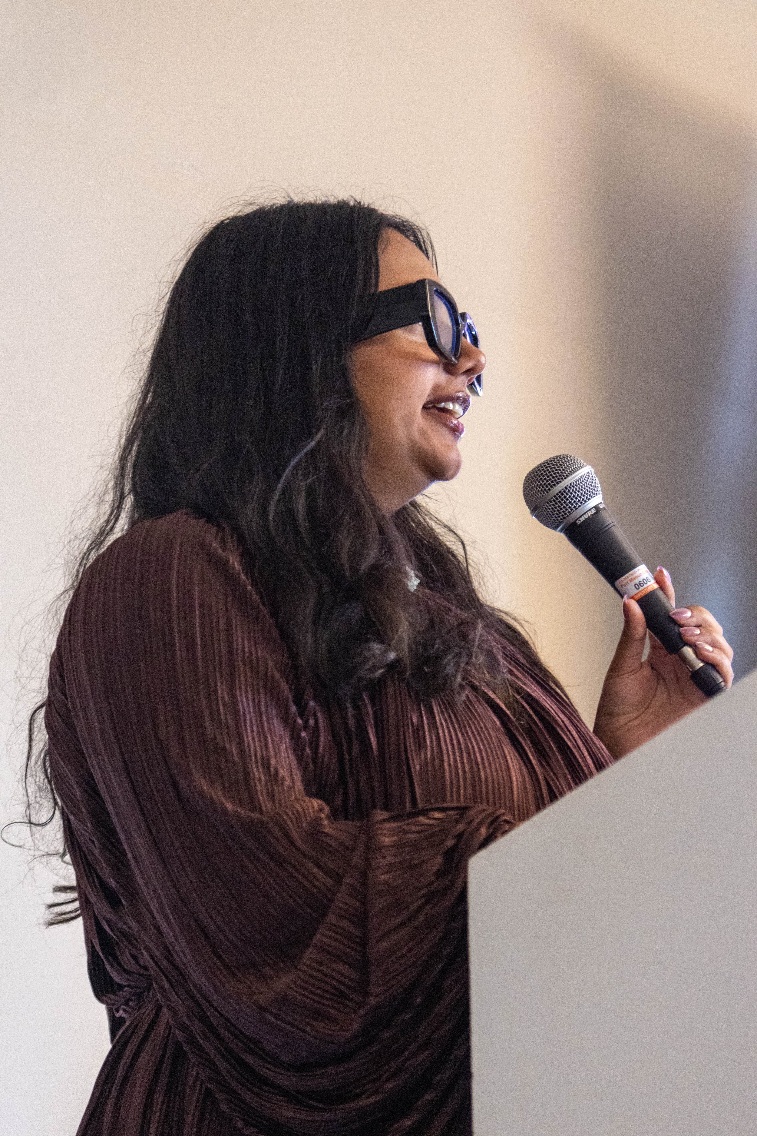 A woman with long black hair, wearing glasses and a maroon striped silky blouse, is speaking into a microphone.