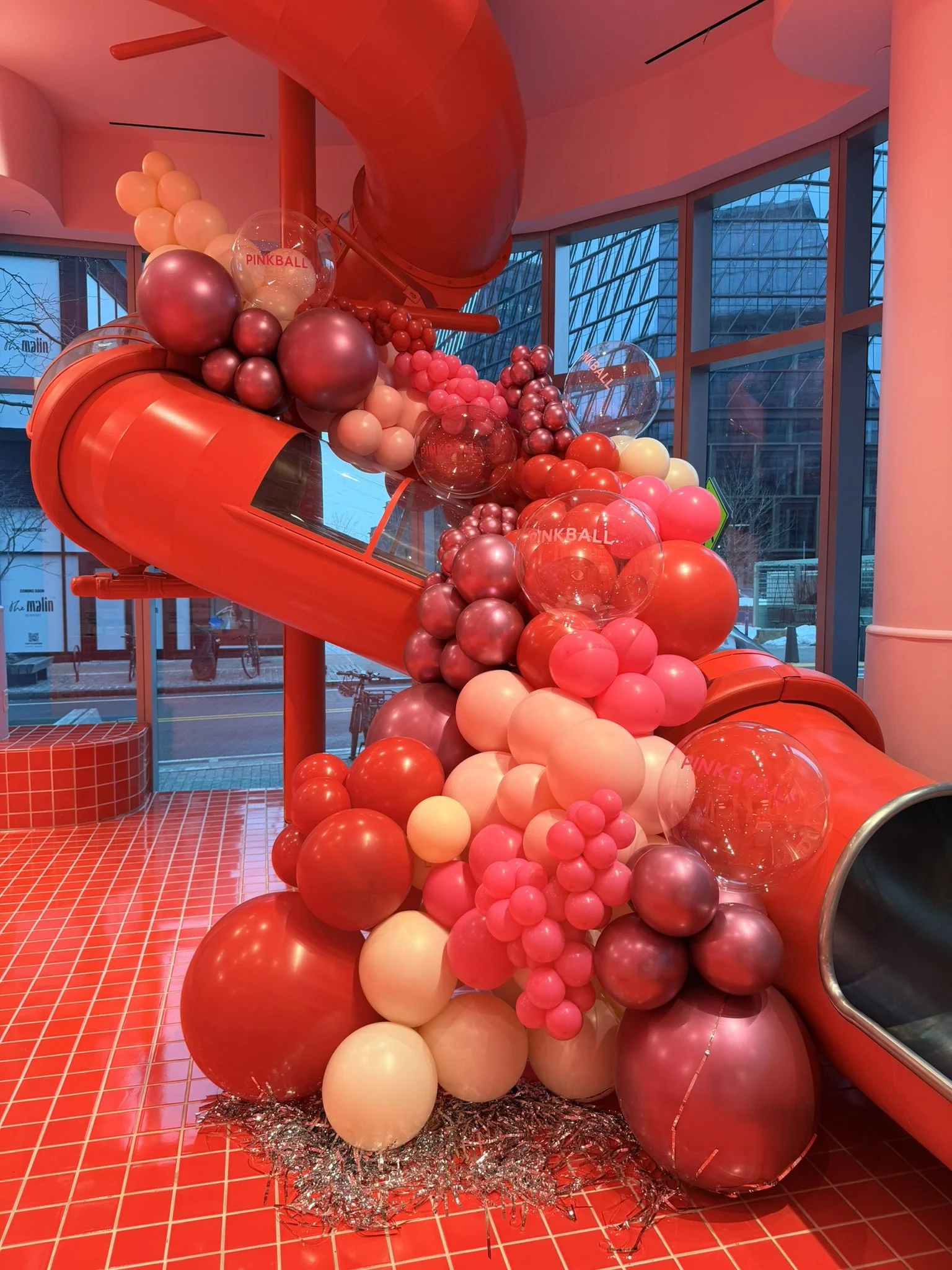 Pinkball event decor with a custom balloon garland in pink, red, cream, gold, and purple around a slide in a modern Boston event space.