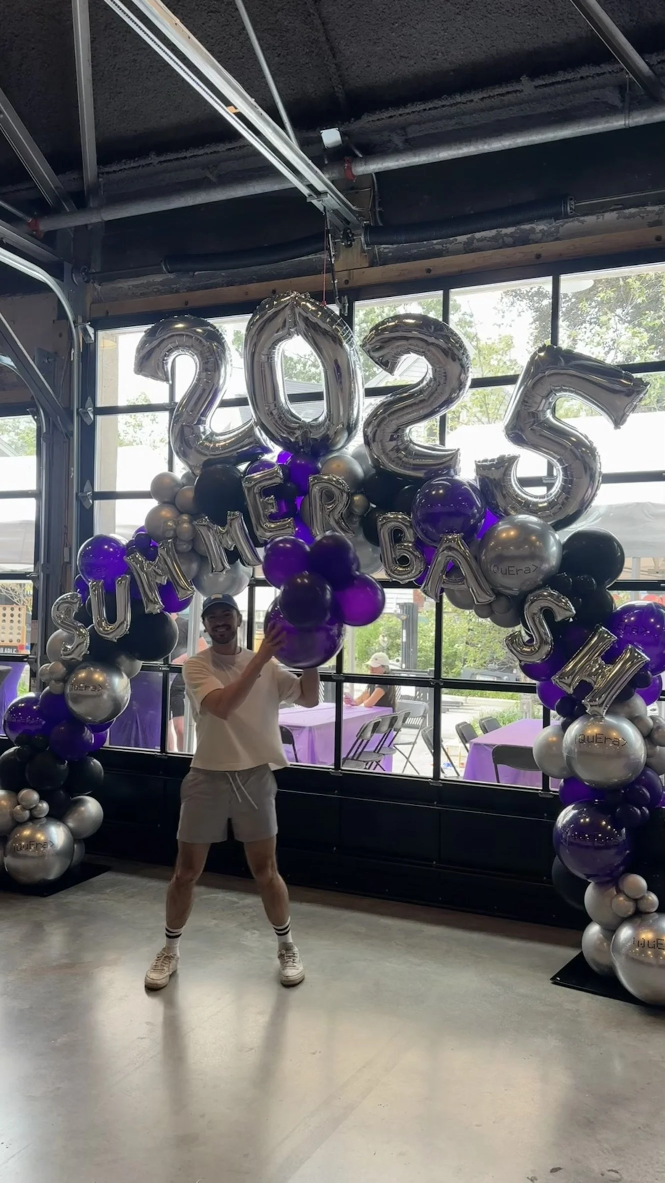 Person standing under a balloon arch decoration with large silver balloons spelling '2022' and smaller balloons in purple, black, and silver. The person is smiling, wearing a white t-shirt, shorts, and sneakers.