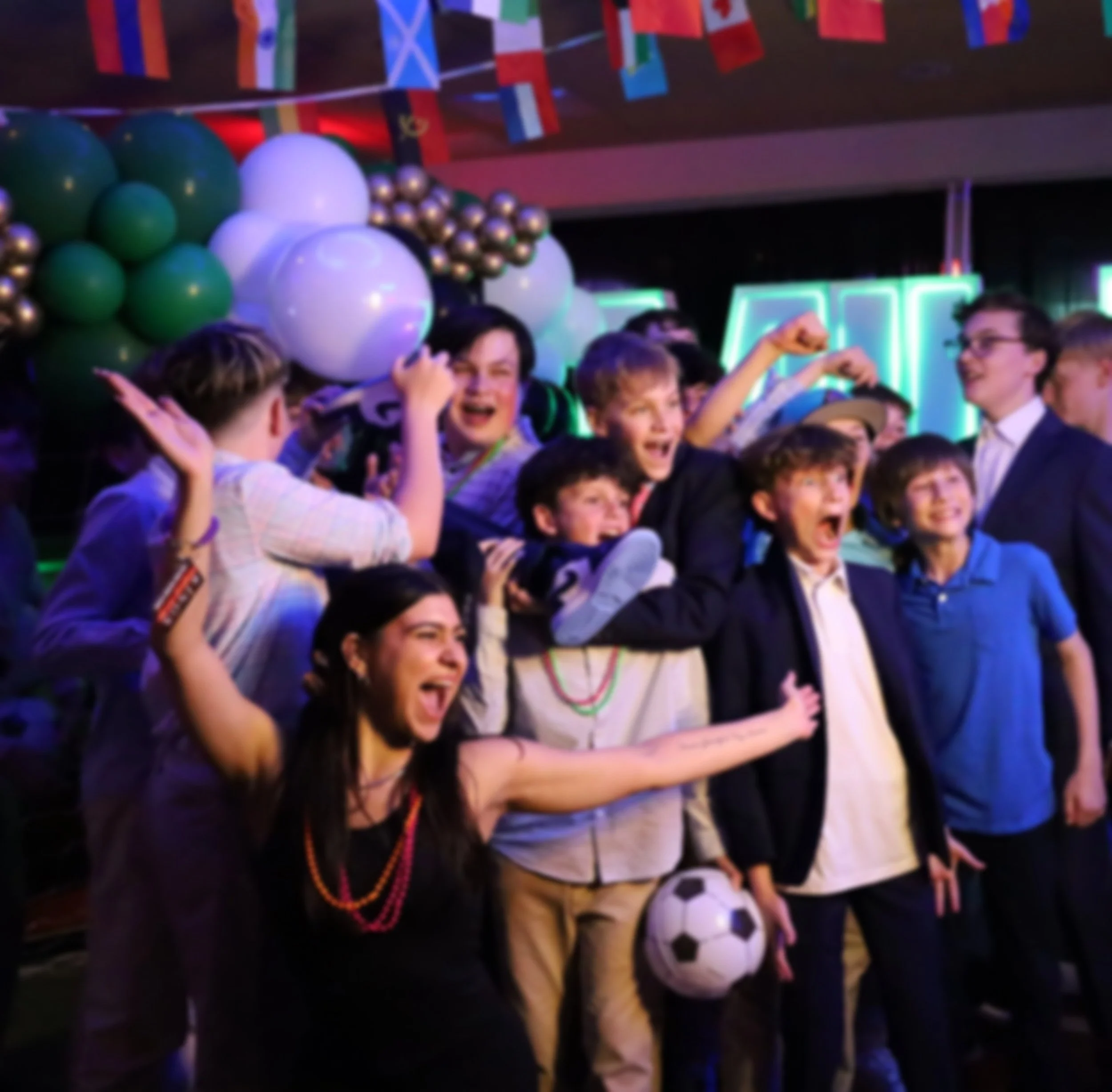 Group of children and teenagers celebrating indoors with balloons and flags, holding and playing with soccer balls.