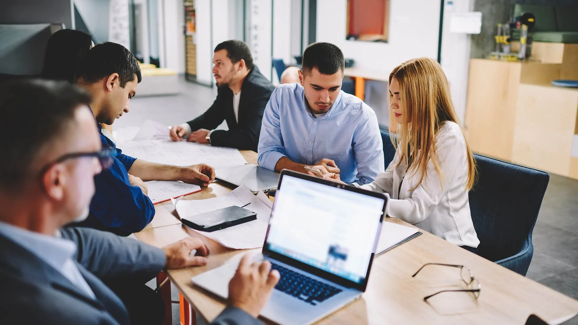 Group of five diverse people working at a long wooden table in a modern office, with laptops and papers, engaged in discussion.