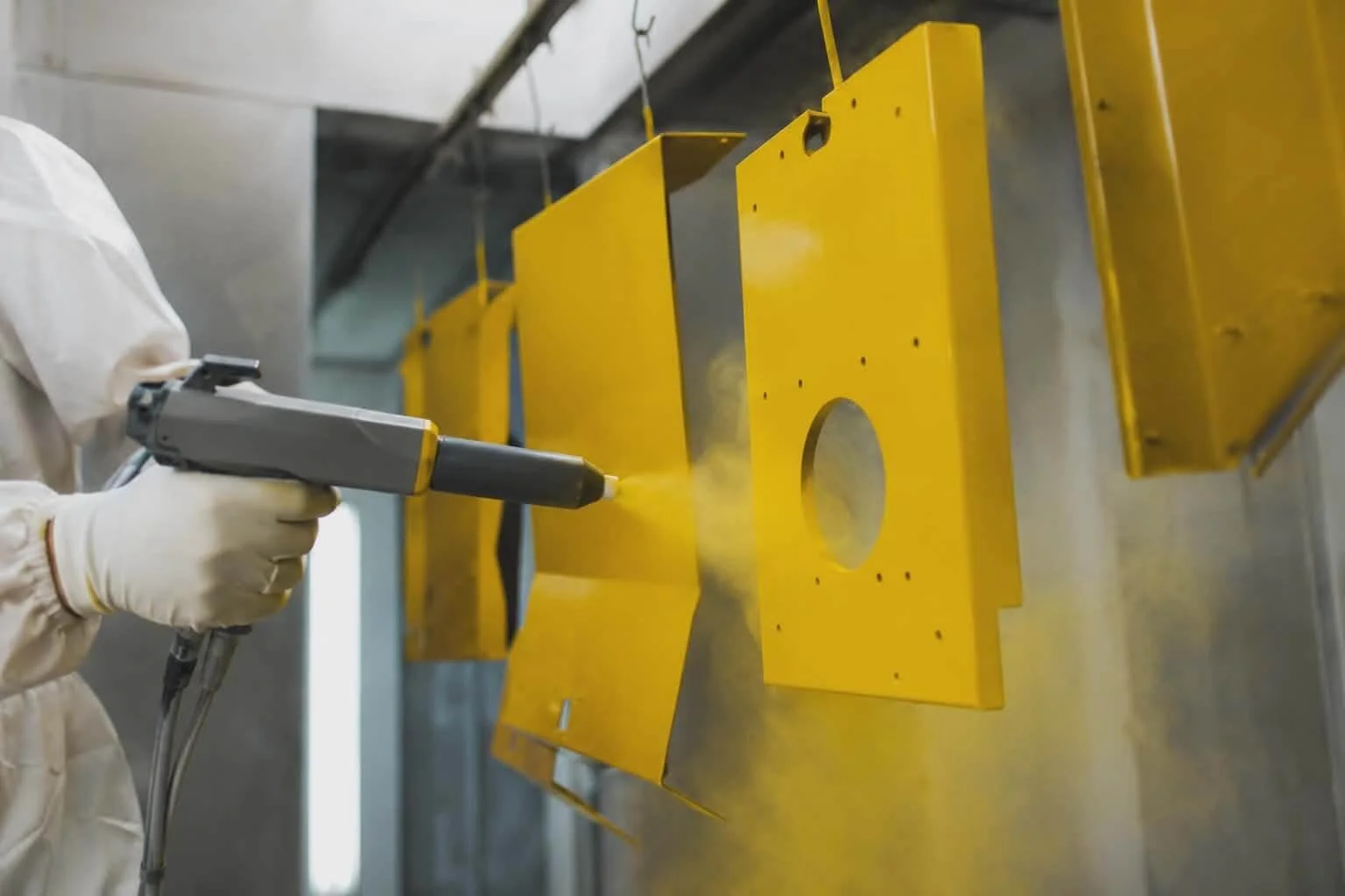 A person in protective gloves uses a spray gun to paint yellow metal parts hanging on a rack in a manufacturing or industrial setting.