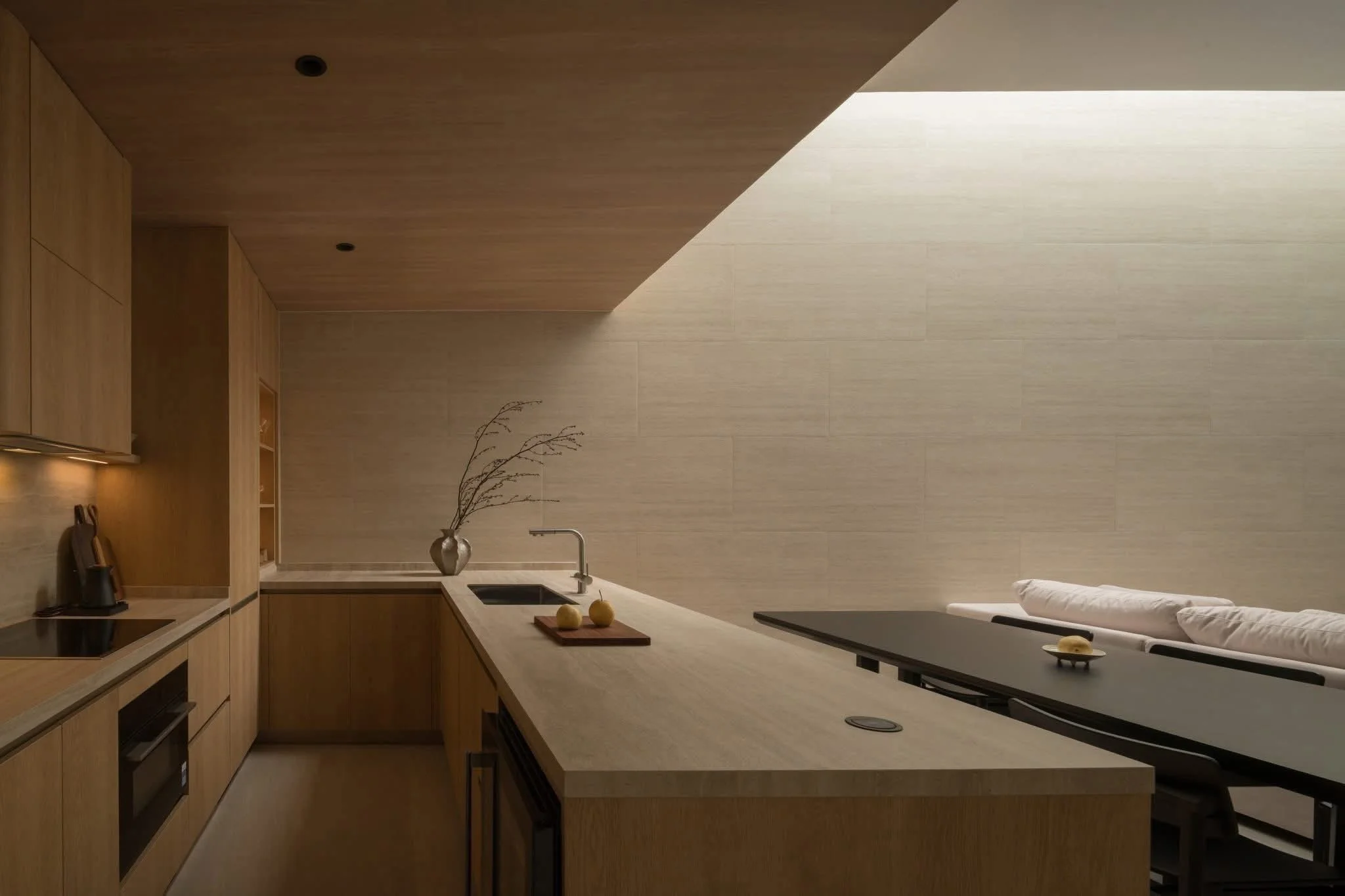 Modern kitchen with light wood cabinetry, beige countertop, and a backsplash. There is a black sink with a silver faucet, two yellow apples on a wooden tray, and a minimalist vase with dried branches. The room features soft lighting and a neutral color palette.