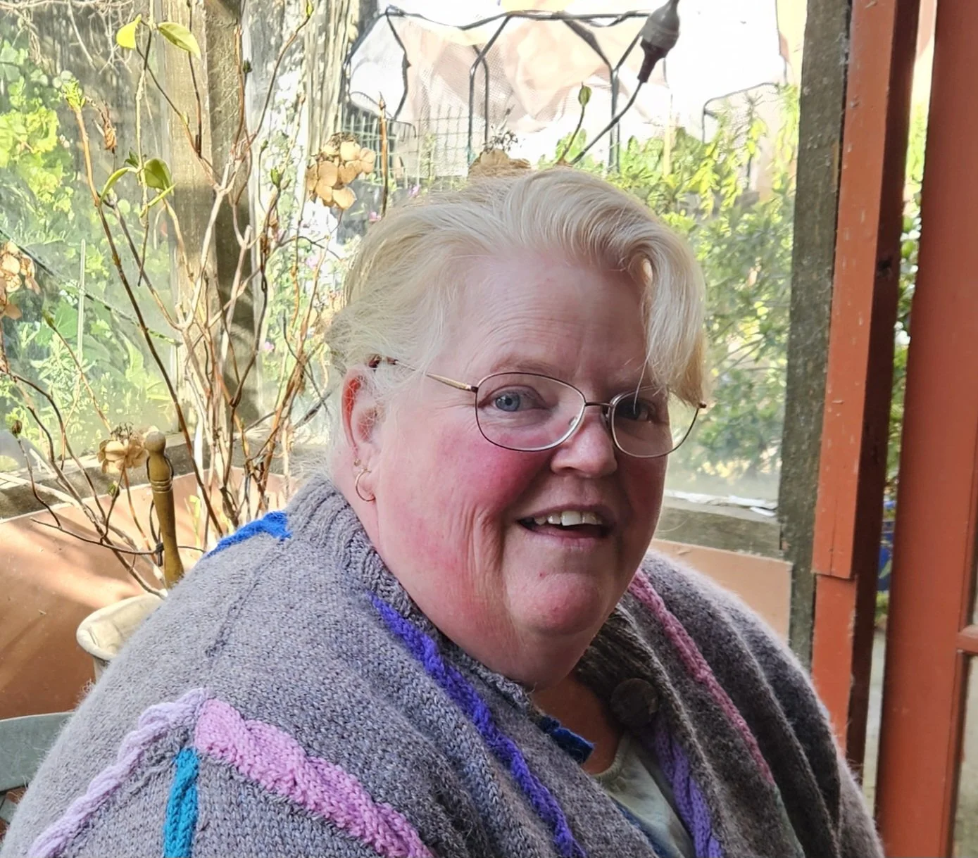 A smiling elderly woman with glasses, sitting outdoors, wrapped in a colorful knitted shawl, with a garden background.
