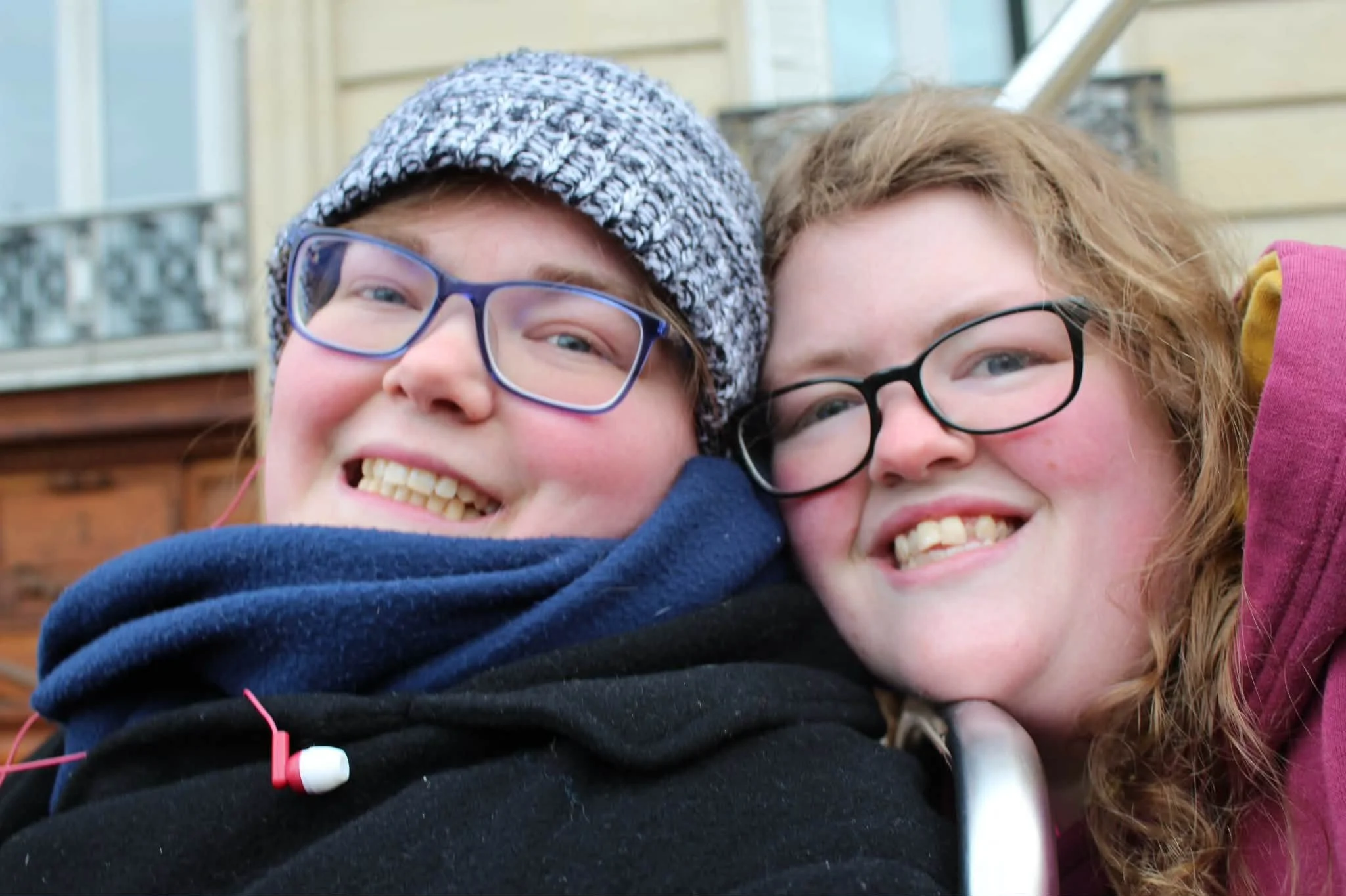 Two teenagers, a boy with glasses and a girl with glasses, are smiling and hugging each other. They are outdoors in front of a house with a porch.